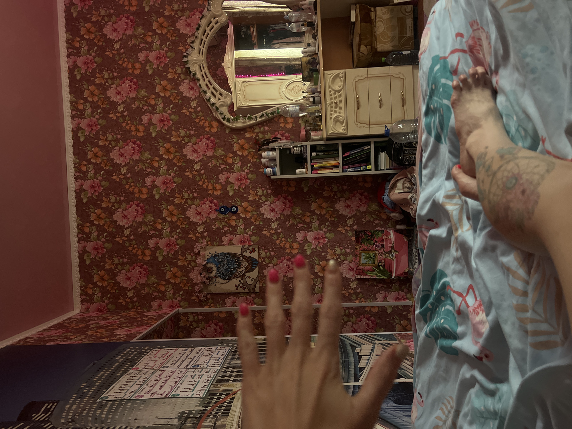 The image captures a bedroom scene with floral wallpaper and a pink ceiling. On the left, a portion of a wall mural depicting a cityscape is visible. In the foreground, a person's hand with painted fingernails is blurred as it is held up in front of the camera, obscuring part of the view. Below, a bed is covered with a duvet featuring a tropical print with flamingos and palm leaves. A person's tattooed leg and foot are visible on the bed. In the background, a vanity with a large ornate mirror stands against the floral wallpaper. A bookshelf filled with books and bottles is to the left of the vanity, and boxes are stacked to its right. A framed picture of an owl hangs on the wall above the bookshelf. The overall impression is a cluttered, personal space.