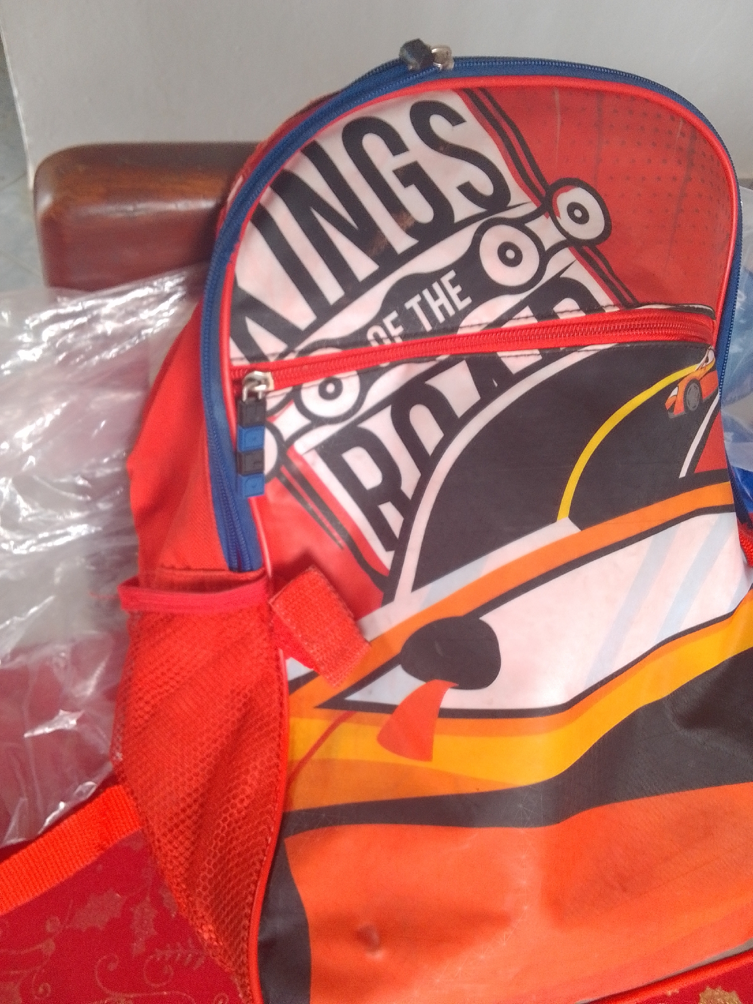 The image shows a close-up of a red backpack with a "KING OF THE ROAD" graphic. The graphic features large black letters on a white and red background, with a stylized race car and road design. The backpack has a zippered compartment, a mesh side pocket, and a blue trim. It appears to be a child's backpack.
The setting is indoors, possibly a home, as indicated by the presence of a wooden armrest and a plastic-covered surface in the background. There are no people visible in the image. The lighting suggests it is daytime. There are no obvious weather cues. The location context provided is La Tagua, Venezuela.
The visible text on the backpack reads "KING OF THE ROAD".