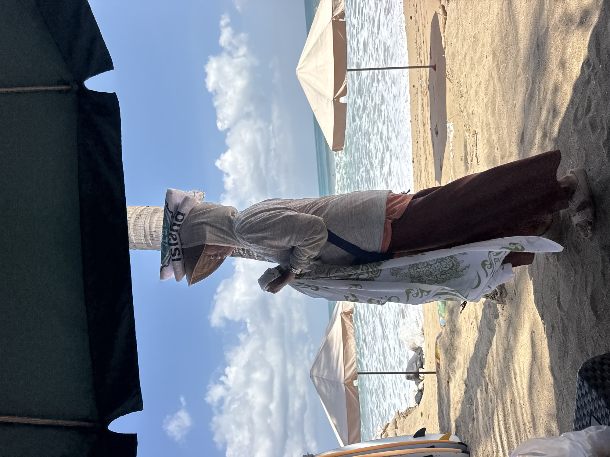 A person wearing a hooded grey jacket, a wide-brimmed hat, and brown pants stands on a sandy beach in Kuta, Indonesia. The person is holding a white towel with a green turtle pattern. The towel is draped over their arm, and they appear to be holding it with their hands. The person's face is partially obscured by the hat and hood. The person is standing under a dark green beach umbrella, and a palm tree is visible behind them. In the background, the ocean is visible with waves breaking on the shore, and two beige beach umbrellas are set up on the sand. Several surfboards are leaning against a sandy embankment to the left of the frame. The sky is blue with scattered white clouds. The ground is covered in sand, and shadows from the umbrella are cast on the sand.