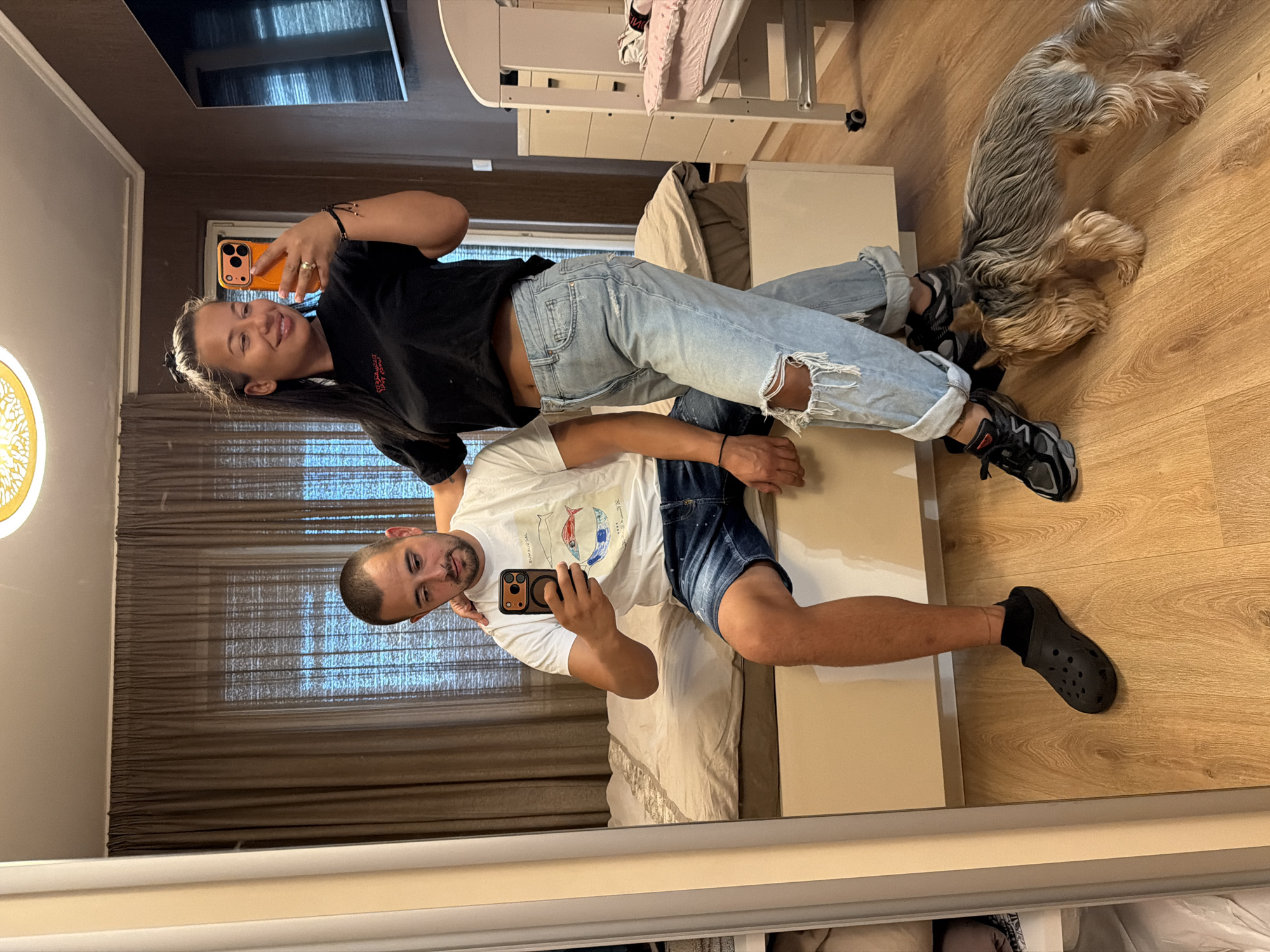 In a room with wooden floors and brown curtains, a man and a woman are taking a mirror selfie. The man, wearing a white t-shirt and blue denim shorts, sits on the edge of a bed. He holds a smartphone, capturing the reflection. The woman, dressed in a black t-shirt and ripped light-wash jeans, stands beside him, also holding a smartphone. A small Yorkshire terrier dog is sniffing the floor near their feet. In the background, a crib and a chest of drawers are visible. The room appears to be in a residential setting within the city of Burgas, Bulgaria.