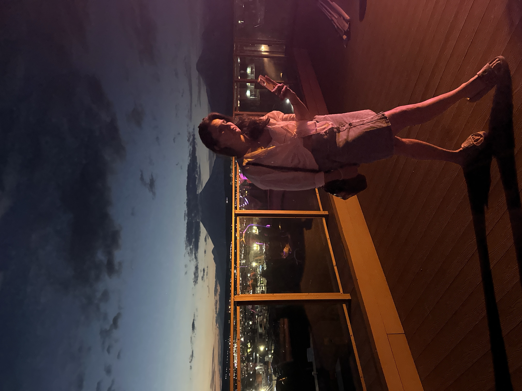 A young woman stands on a wooden deck at dusk, holding a smartphone. She is wearing a light-colored blouse and a denim skirt, with leopard-print slip-on shoes. A crossbody bag is slung over her shoulder. In the background, a city skyline is visible with lights twinkling, set against a twilight sky. Mountains are silhouetted in the distance. The scene appears to be a rooftop bar or observation deck in Petropavlovsk-Kamchatsky, Russia, given the presence of the distinctive mountains known as the Avachinsky and Koryaksky volcanoes. The lighting suggests the setting is illuminated by artificial lights, casting a warm glow on the foreground.