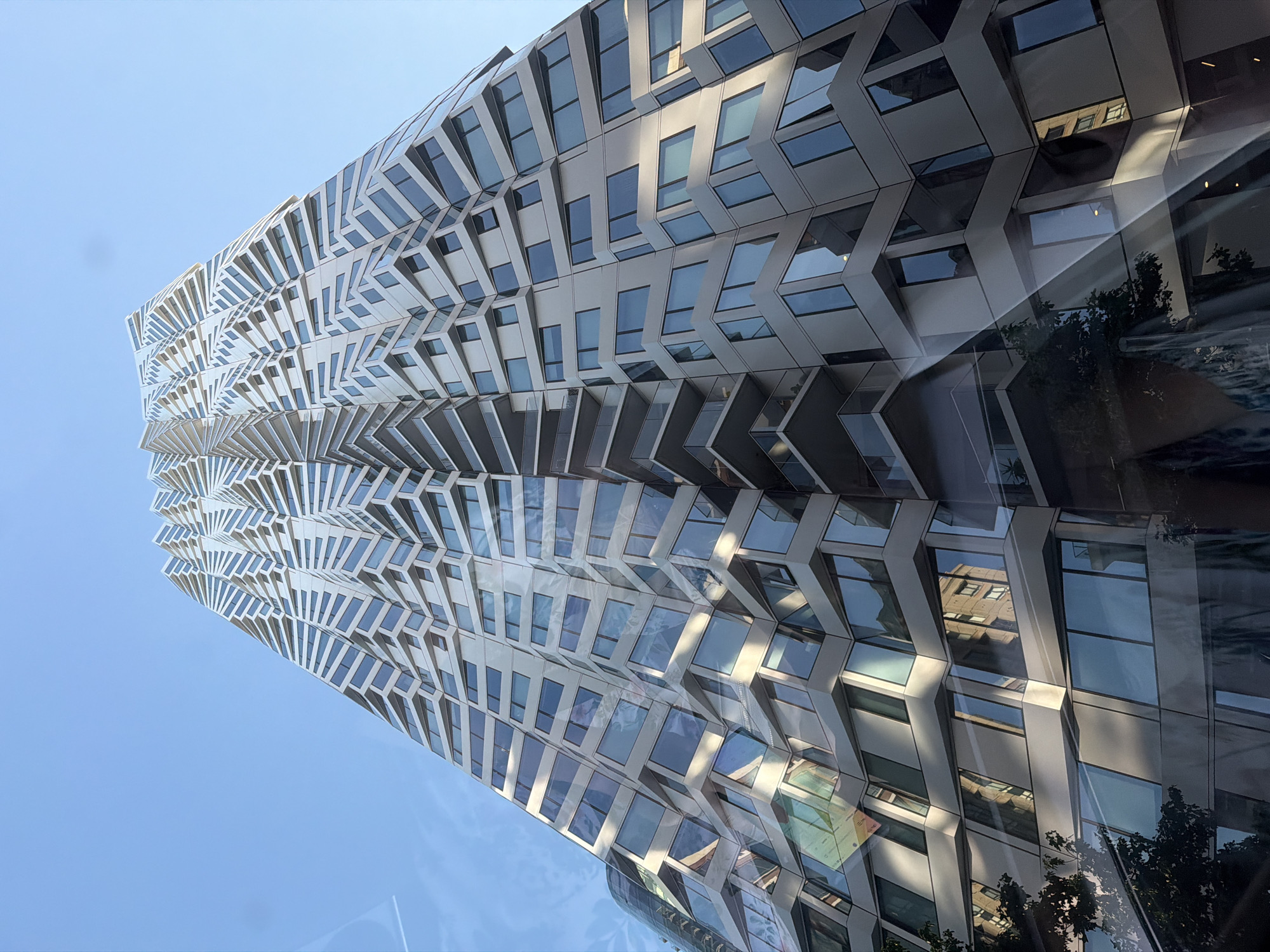 This is a low-angle, eye-level shot of the exterior of a modern skyscraper in San Francisco, California, USA. The building's facade is composed of numerous irregularly shaped panels that create a complex, geometric pattern, giving it a deconstructed, almost fragmented appearance. The panels are a light color, possibly white or a light gray, and they are interspersed with large glass windows that reflect the surrounding environment. The sky above is a clear, bright blue. The photograph appears to have been taken during the daytime, as evidenced by the strong sunlight and the clear visibility of the building's details. The reflections on the glass windows show other buildings and trees, suggesting an urban setting. The camera angle emphasizes the height and unique architectural design of the skyscraper.
