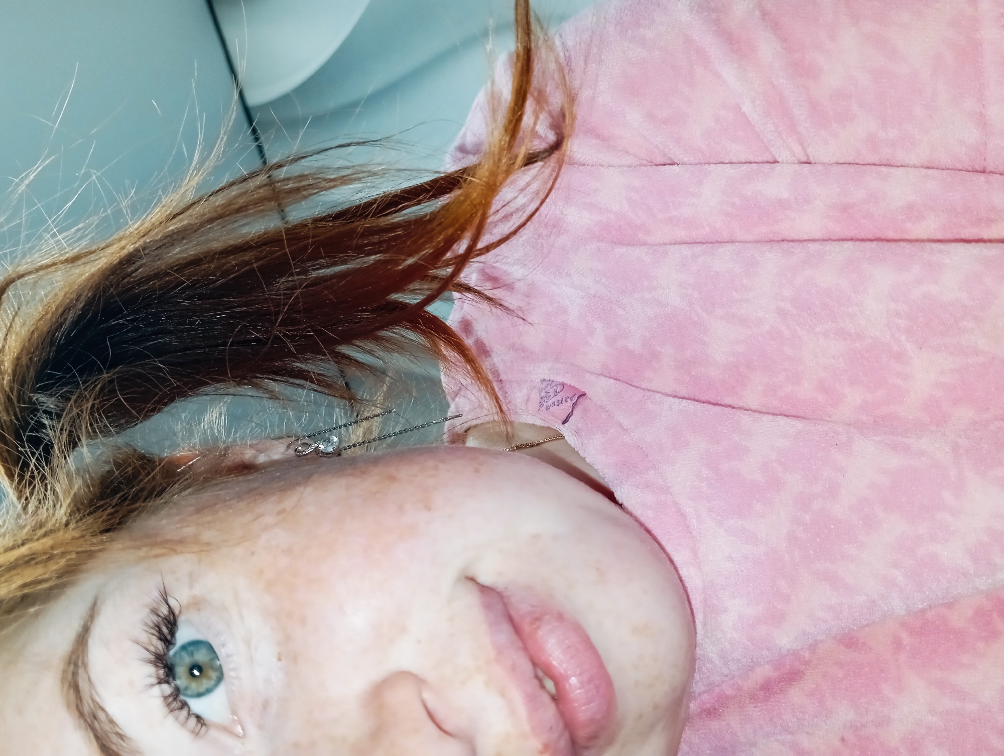 A close-up, angled view shows a person with fair skin and freckles, possibly female, wearing a pink patterned garment, perhaps a bathrobe or blanket. They have reddish-brown hair and a light blue eye is visible. The background includes a white surface and a section of a light blue wall or surface. No information connects this image to the city of Yurgamysh, Russia.