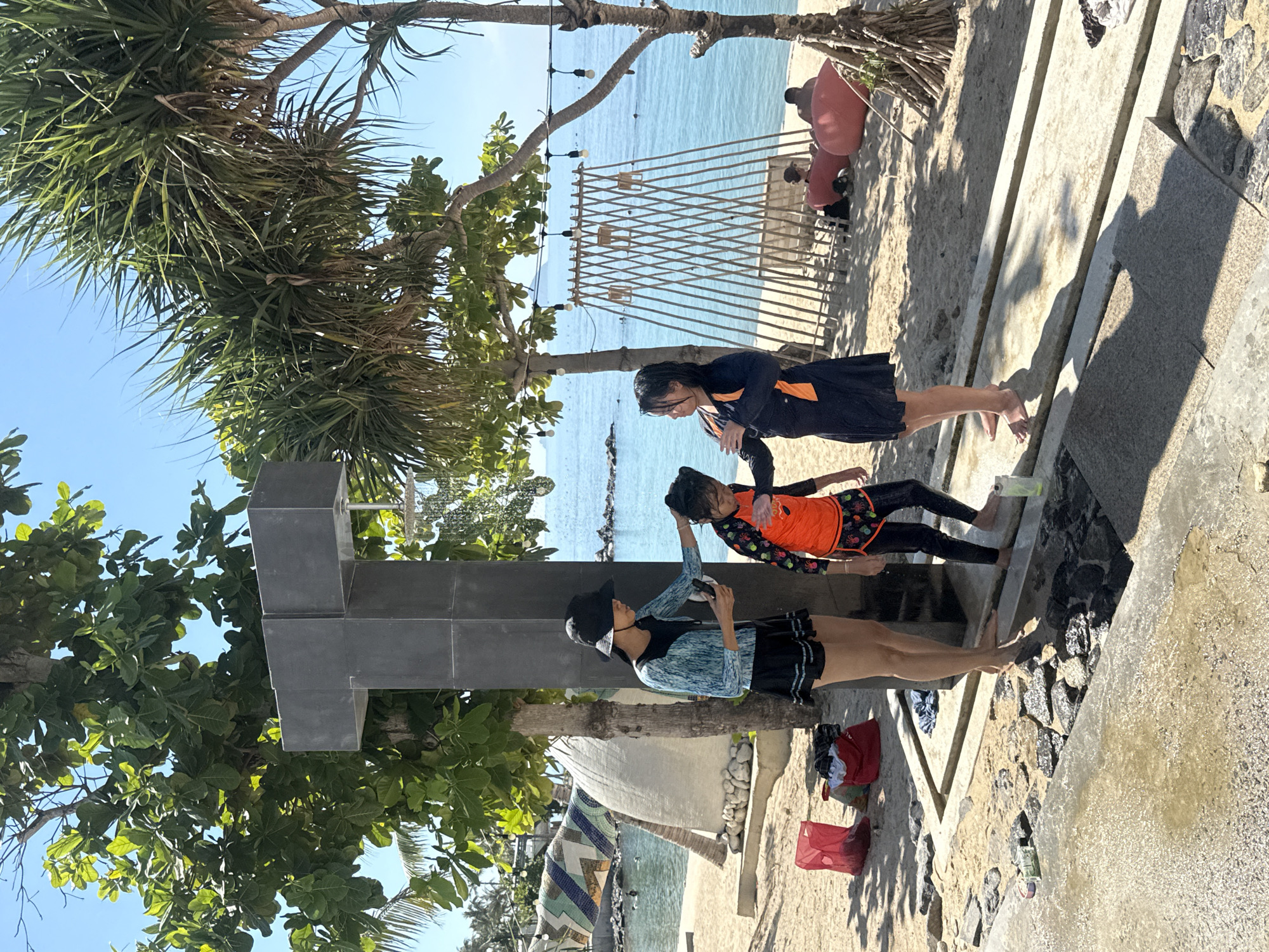 Three young females, appearing to be children, are positioned on a sandy beach near a concrete structure with an overhead shower. The female on the left wears a black hat, a blue and white patterned long-sleeved rash guard, and a black skirt. She holds a mobile phone in her right hand and appears to be interacting with the middle female. The middle female, wearing an orange and black patterned rash guard with black shorts, looks towards the female on the left. The female on the right is dressed in a black long-sleeved rash guard with orange accents and a black skirt, facing towards the left. A showerhead is visible above and to the right of the females. In the background, the ocean is calm with a clear blue sky. Several palm trees and other tropical vegetation are present on the beach. Red bags and other items are scattered on the sand to the left of the group. Further back, a white, wave-like structure with colorful mosaic tiles is visible. A bamboo-like structure with lights strung between poles stands on the sand near the water. Two individuals are seated on large red beanbag chairs in the background. The scene suggests a beach resort or public beach area, potentially in Bac Nha Trang Ward, Vietnam, given the tropical environment.