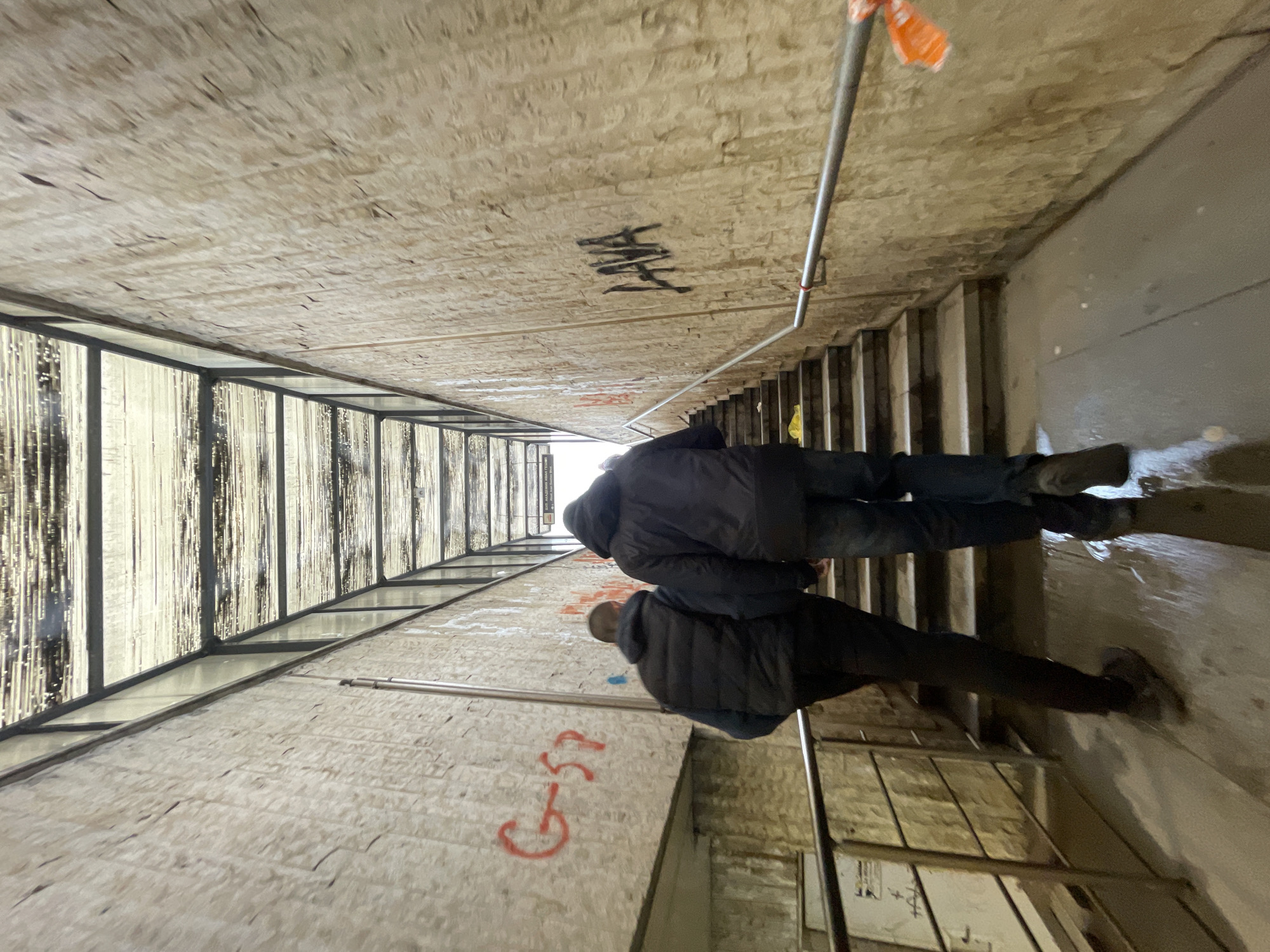 Two people descend a dimly lit, narrow stairway within an enclosed, pedestrian underpass. The underpass has a textured, concrete ceiling and walls marked with graffiti. A metal railing runs along the right side of the stairs. A windowed structure is on the left. The underpass appears damp. The location is likely within Sofia, Bulgaria, based on graffiti styles and underpass architecture common to the city.