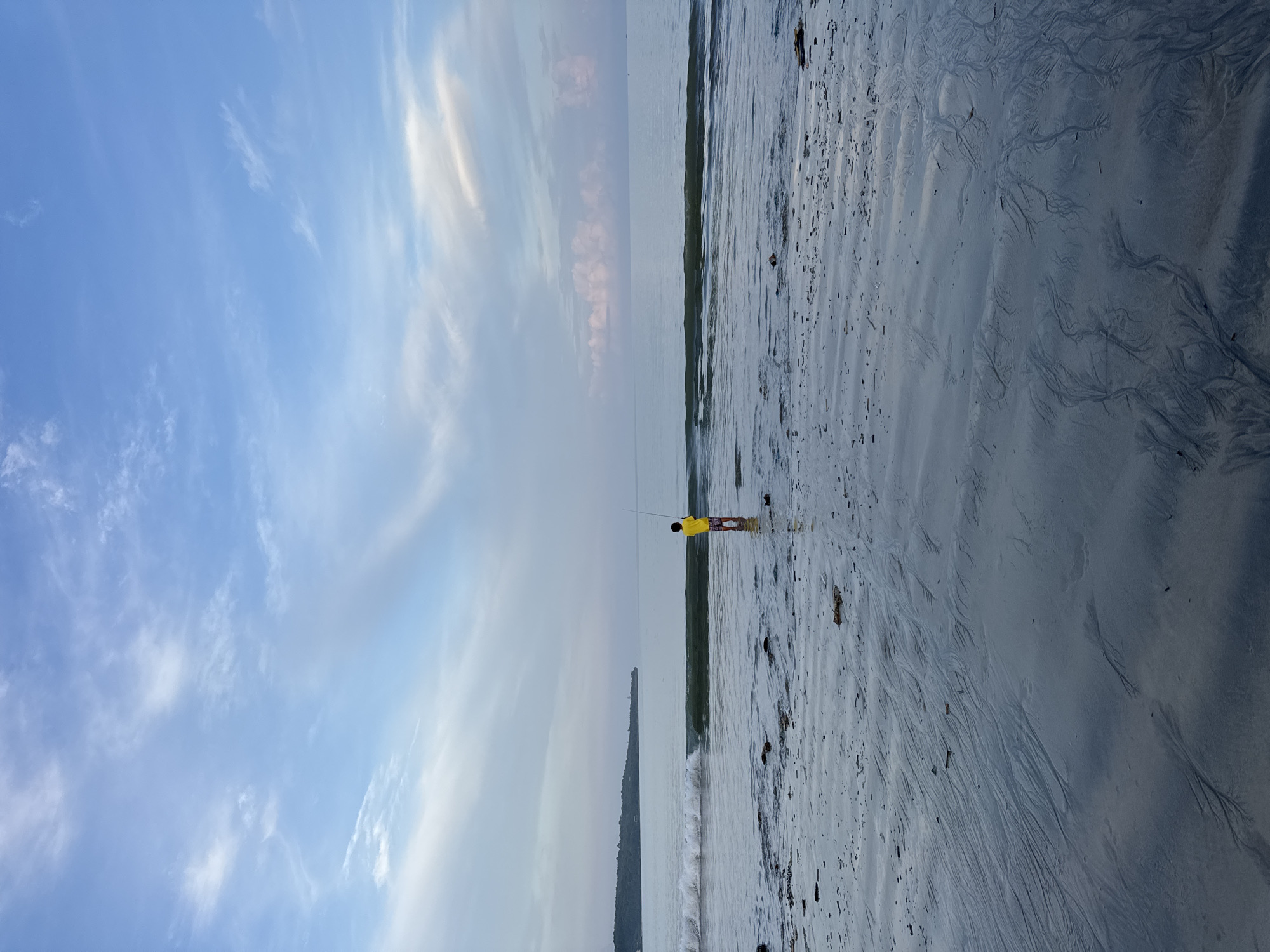 A vertical eye-level shot of Jimbaran beach, Indonesia, shows a person fishing in shallow water, facing the sea. The sky is blue with streaks of white clouds. The beach slopes gently down to the sea with patterns on the sand. A wave is breaking along the shore.