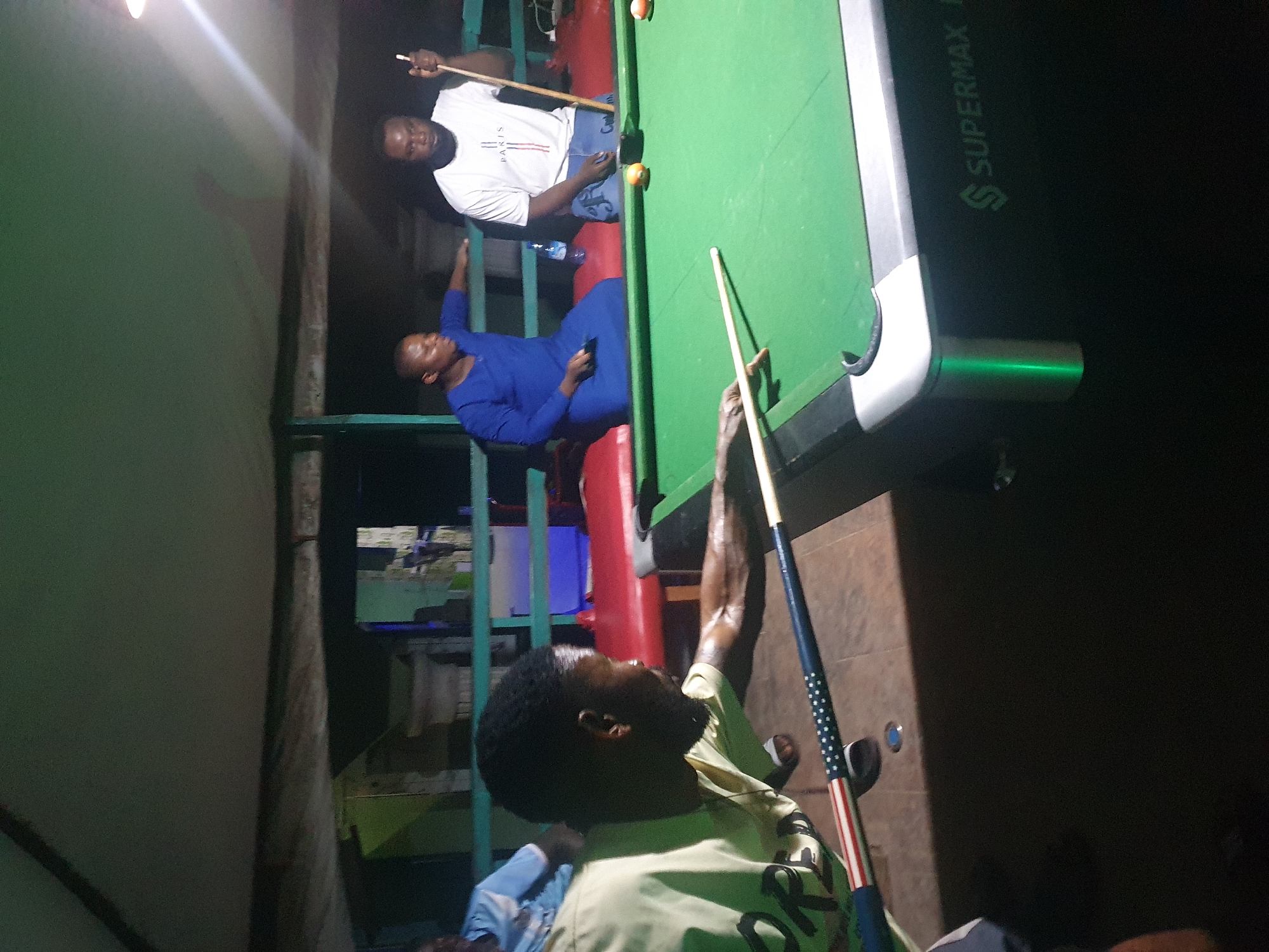 The image depicts an indoor setting, likely a bar or recreational center, in Lokoja, Nigeria, where four men are engaged in a game of pool. A green pool table dominates the foreground, with several balls visible on its surface. One man, wearing a light-colored t-shirt with a U.S. flag design on his cue stick, is in a striking pose, reaching to hit a ball. Another man, in a blue robe-like garment, is seated, holding a smartphone. A third man, dressed in a white t-shirt and blue shorts, stands by the table, holding a cue stick. A fourth man, partially visible in the background, wears a light blue shirt. The environment appears somewhat dimly lit, with artificial lighting from above. A water bottle is present on a red surface behind the pool table. The presence of a "SUPERMAX" logo on the side of the pool table suggests it is a branded piece of equipment. The overall atmosphere suggests a casual social gathering focused on the game.