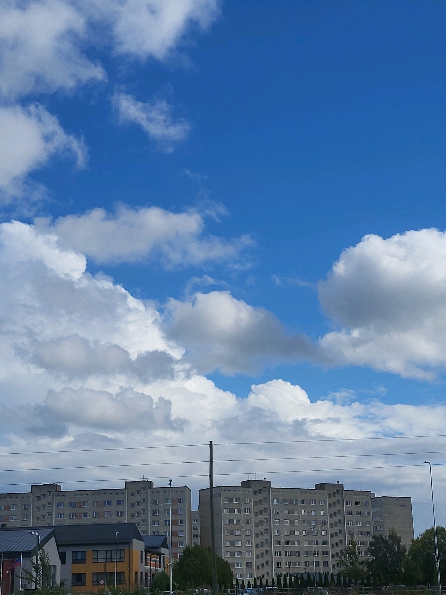 The image depicts a section of the city of Jelgava, Latvia, characterized by a bright blue sky filled with puffy white clouds. In the foreground, a series of large, multi-story residential buildings, typical of Soviet-era architecture, stretch across the frame. These buildings are uniform in their gray facade and feature numerous windows. Power lines run horizontally across the scene, intersecting the view of the buildings. Below the main residential blocks, there are smaller, modern structures with colorful accents. Trees and streetlights are also visible, suggesting an urban environment. The overall impression is one of a daytime scene in a developed urban area.
