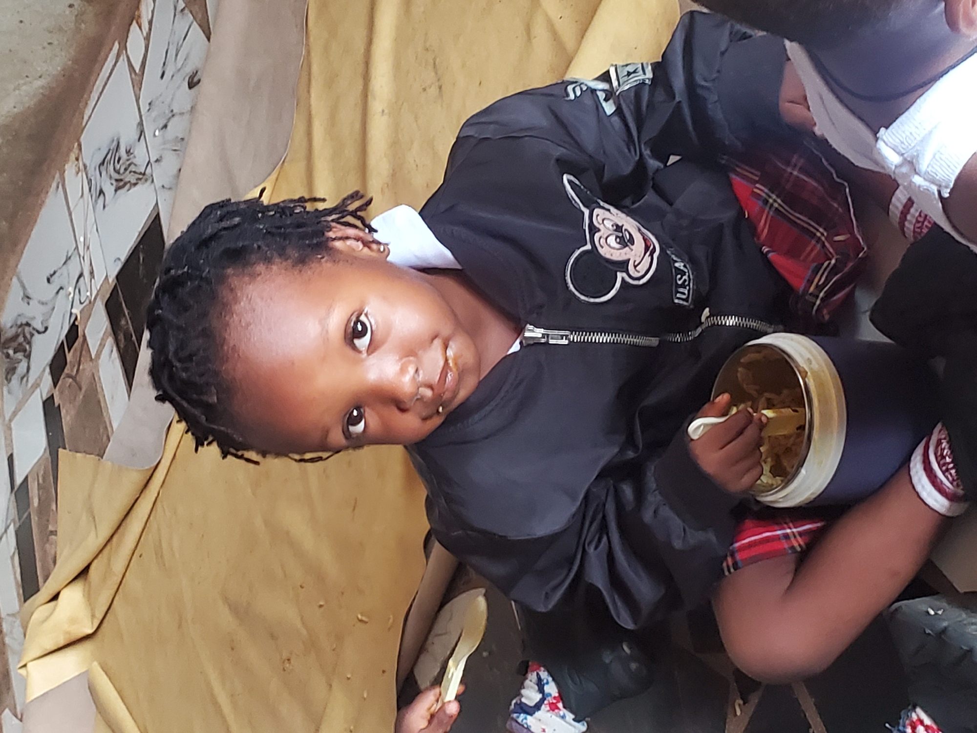 A young Black child with short dreadlocks is seated indoors, holding a bowl of food with a spoon. She is wearing a dark jacket with a Mickey Mouse patch and a white collared shirt underneath. She is looking at the camera. A portion of another child's head, wearing a white shirt and red plaid, is visible to the right. The setting appears to be a simple interior space. The location is stated to be Ukwu-Abwa, Nigeria.