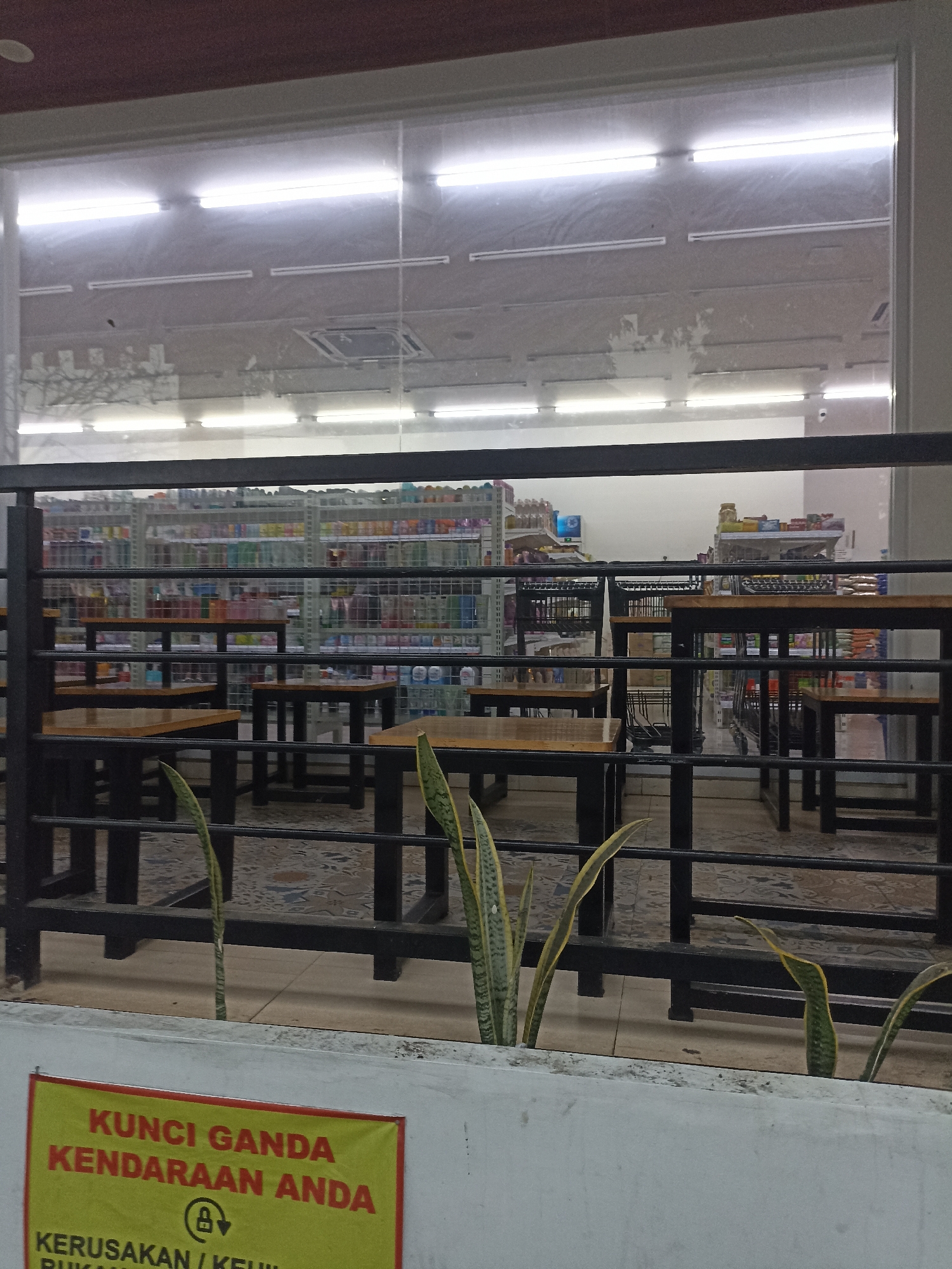 In Tompokersari, Indonesia, the image depicts an outdoor seating area with multiple wooden tables and black metal chairs set up in front of a convenience store visible through a large glass window. The store's interior is well-lit with fluorescent lights and stocked with shelves of various products. In the foreground, a yellow sign with red lettering reads "KUNCI GANDA KENDARAAN ANDA" (Double Lock Your Vehicle) and includes a symbol of a padlock with an arrow. Snake plants are visible growing in a concrete planter in front of the seating area. The overall scene suggests a public space designed for customers to relax or wait while at the convenience store.