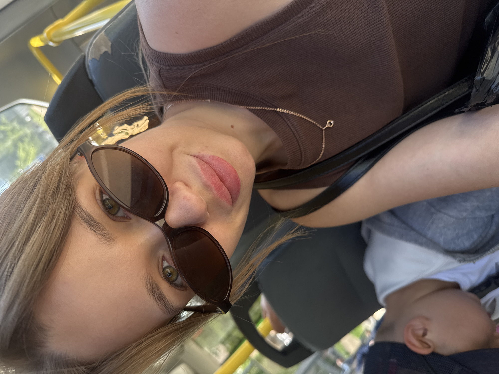 A young woman with blonde hair and sunglasses is taking a selfie on a bus, possibly in Chaikovsky, Russia, given the presence of a sleeping child behind her. She is wearing a brown sleeveless top and a delicate necklace. The bus interior is visible with yellow handrails and dark seating. The child appears to be wearing a gray hoodie and a black baseball cap.