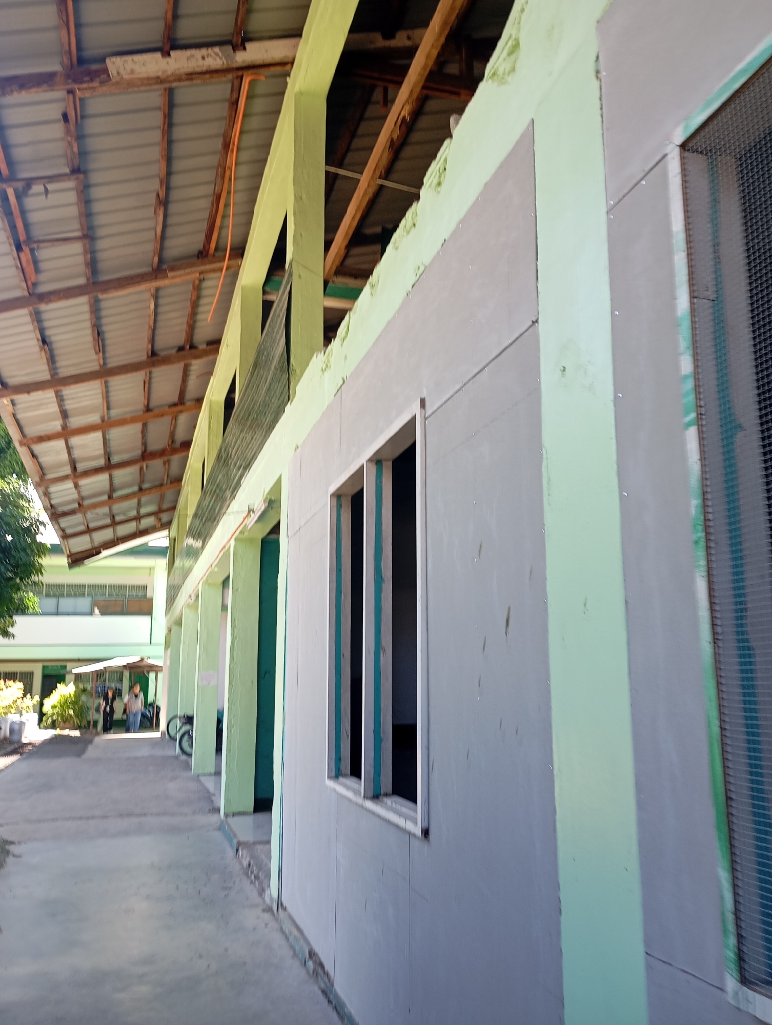 The image depicts an exterior view of a building, likely under construction or renovation, located in Davao City, Philippines. The building features a light green painted facade with grey panels installed on the walls. A prominent element is a three-pane window on the grey paneling. The roof structure, made of corrugated metal and wooden beams, is visible in the background, suggesting an open-air walkway or covered area. Further down the walkway, two individuals are walking, and a motorcycle is parked. The overall environment suggests a school or institutional setting, given the linear arrangement of buildings and the open courtyard area. The lighting indicates it is daytime.