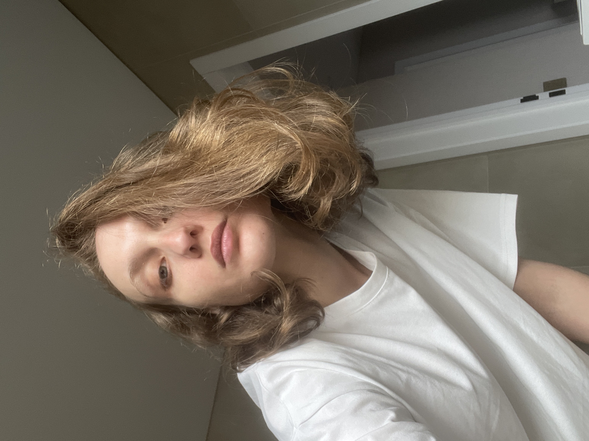 The image is a head-and-shoulders portrait of a young individual, likely female, with light brown or blonde wavy hair that appears tousled. The person has fair skin, a neutral to slightly pensive expression, and is looking directly into the camera. A small dark mark is visible on the upper lip. They are wearing a plain white t-shirt.
The photograph is taken from a low angle, with the subject's head tilted significantly to the side, suggesting they might be lying down or the camera was held at an unusual angle. The background is a simple, muted light grey or beige wall or ceiling. Above the person's head, a white architectural feature is visible, possibly a window frame, a ceiling vent, or a piece of white furniture like a shelf or cabinet, with some dark lines and shadows indicating recesses or openings.
The lighting is soft and diffused, characteristic of an indoor setting during daytime, with no direct sunlight or harsh shadows. No specific activity is depicted beyond posing for the portrait. The overall scene suggests a calm, unadorned indoor environment in Podshivalovo, Russia. There is no visible text in the image.