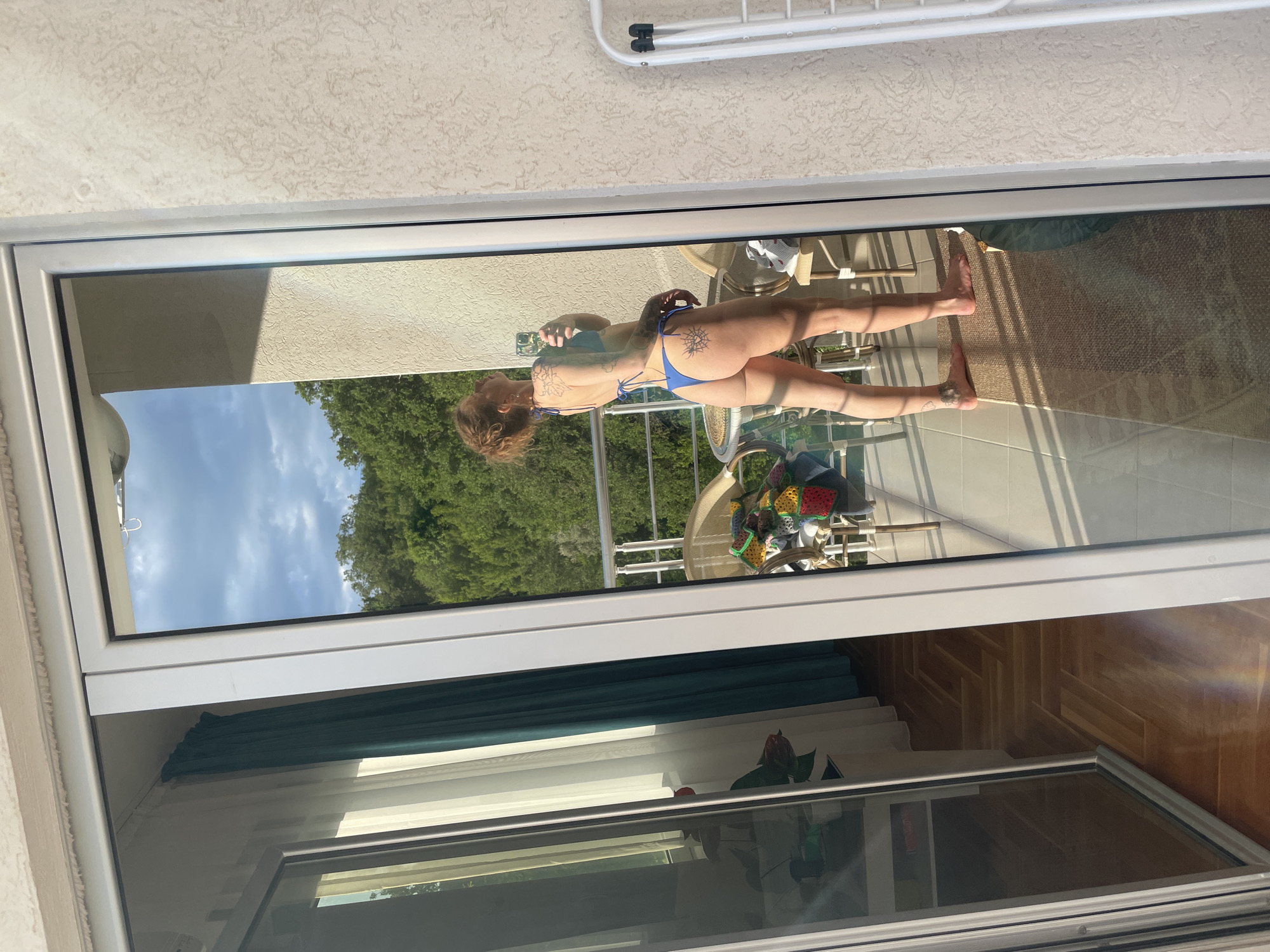 A partially opened window frames the reflection of a woman taking a photo of herself on a balcony. She wears a blue bikini. Her back is to the window. The background outside shows lush green trees under a blue sky with scattered clouds. A clothes drying rack is visible at the top of the frame outside. The balcony contains chairs and a table with an object that may be covered with a colorful crocheted blanket. The room inside the window shows a teal curtain and a wooden floor. The location is likely Rafailovići, Montenegro, based on the architectural style and surrounding landscape.