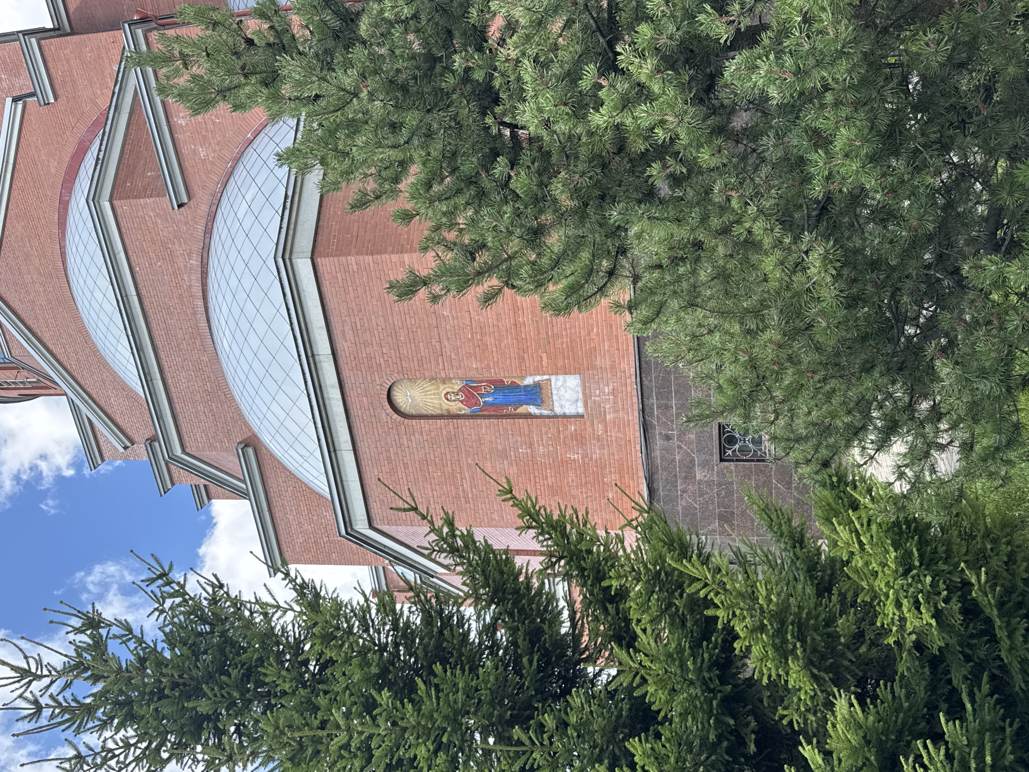 The image depicts a close-up view of a red brick building, likely a church or cathedral, with several domed roofs. A mosaic of a religious figure in blue and red garments against a gold background is visible on the brick wall. The building is partially obscured by the dense green foliage of two conifer trees in the foreground. A small, circular window with decorative metalwork is also visible on the lower part of the building. The sky above is blue with scattered white clouds. The location is identified as Sinyavino, Russia.