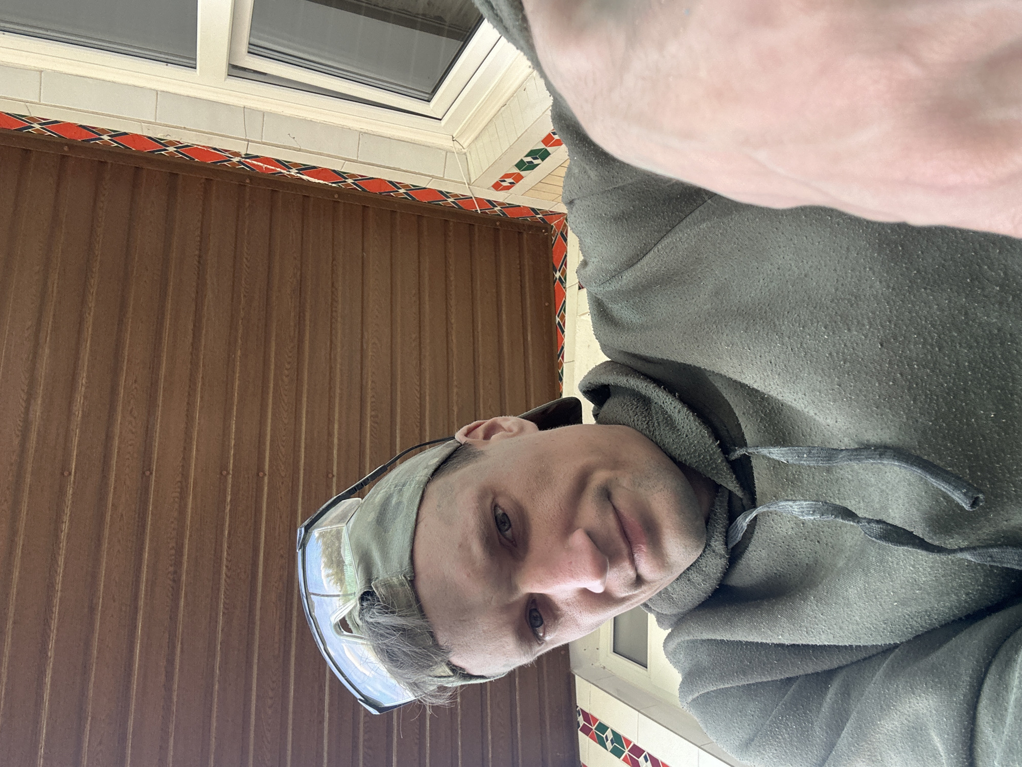 A man is taking a selfie outdoors. He is wearing a green baseball cap with safety glasses pushed up onto it, and a green hooded sweatshirt. He has fair skin and is looking at the camera with a neutral expression. The background shows a brown wooden ceiling, a white window frame, and a decorative tile border with red, green, and black geometric patterns. The image appears to have been taken on a porch or under an overhang. The overall scene suggests a casual, possibly work-related, outdoor setting. There is no specific indication of the city of Makiv, Ukraine, or any other geographical context beyond what is visually present in the image.