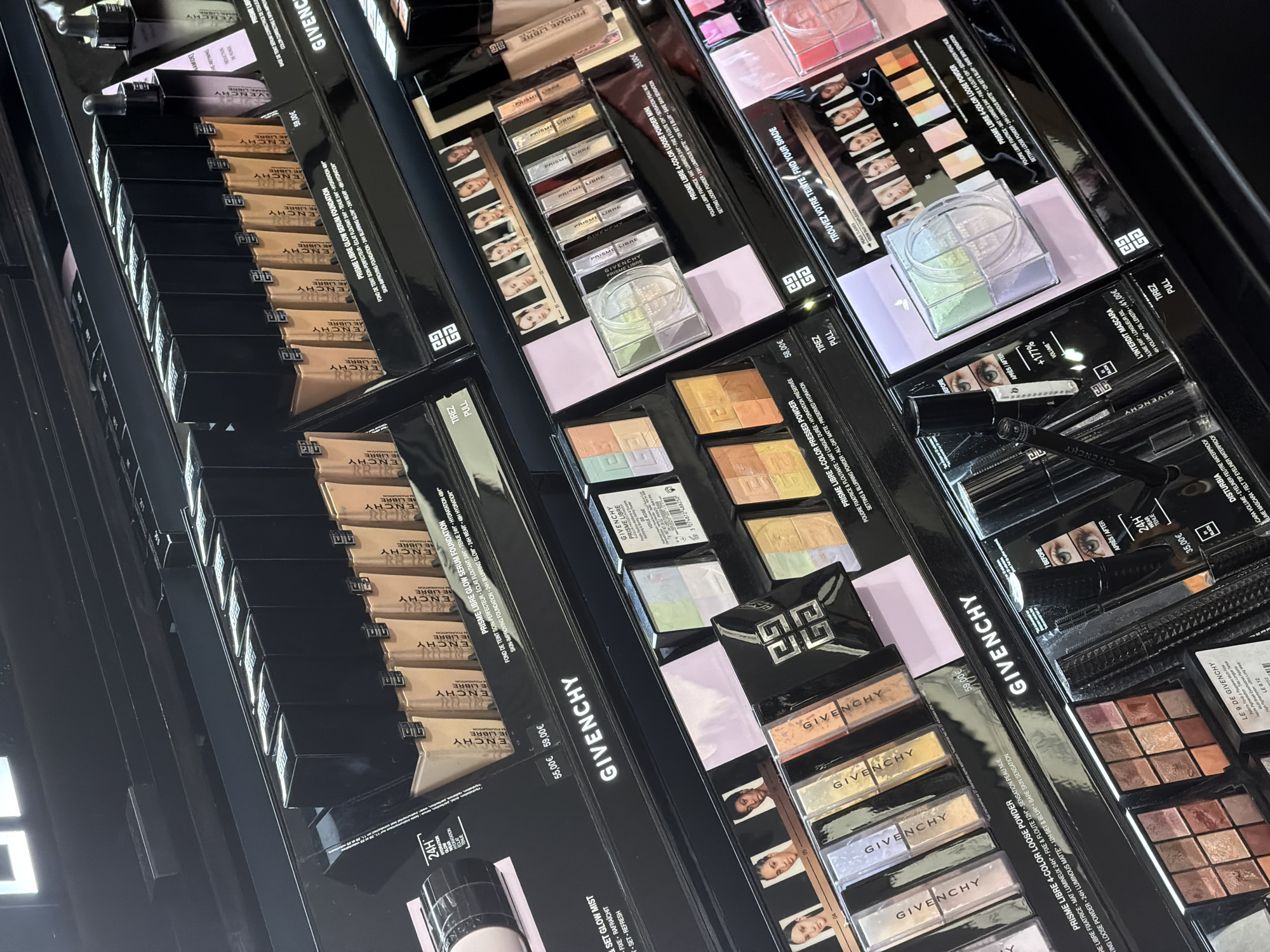 The image is a close-up, angled shot of a brightly lit retail display of Givenchy cosmetics. The display is stocked with various makeup products, including foundation bottles, powder compacts, and eyeshadow palettes. The products are neatly arranged on multiple shelves, with the Givenchy logo prominently visible on several items and the display itself. The scene appears to be an indoor retail environment, likely a department store or a dedicated beauty store. The overall impression is one of a well-organized and upscale cosmetic counter. The question about Barcelona, Spain is not answerable from the provided image.