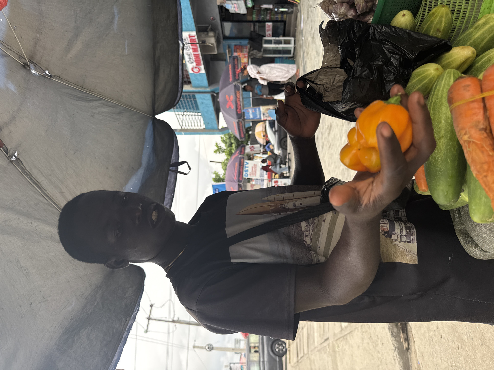 A young Black male stands under a gray tarped canopy, holding an orange bell pepper in his left hand. He wears a black t-shirt with a graphic print of a rocket and buildings. A black strap is slung across his chest. In front of him, a woven basket contains cucumbers and carrots, and a black plastic bag. Behind him, a street scene unfolds with buildings, other vendors with umbrellas, cars, and people. A sign reads "Great Bella MINI MART". The scene appears to be a street market in Ado, Nigeria, under an overcast sky.