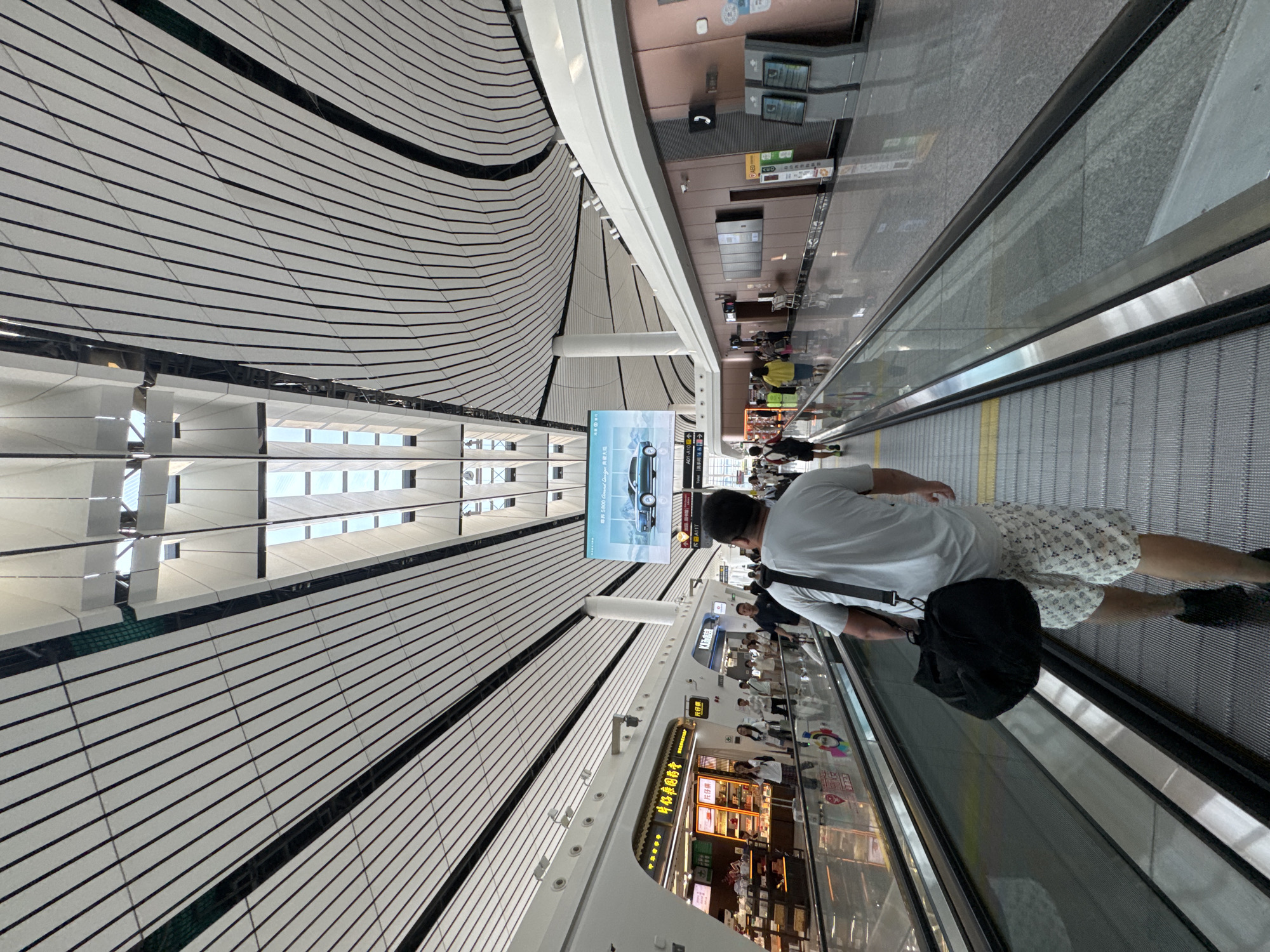 The image depicts an interior scene within the Daxing International Airport in Guangyang District, China. The perspective is from a moving walkway, looking forward and slightly upwards. A man, seen from behind, is walking on the walkway, carrying a black duffel bag over his left shoulder. He is wearing a light-colored t-shirt and patterned shorts.
The environment is characterized by a modern architectural design with a distinctive undulating ceiling constructed from white panels and black support beams, interspersed with skylights that allow natural light to filter in. On either side of the walkway are shops and service areas. To the left, a storefront with illuminated displays shows products, and people are seen browsing. To the right, a wall panel features a large digital advertisement displaying a blue car. Further down the walkway, other individuals are visible, some walking and others standing. The floor is highly reflective, suggesting a polished surface. The overall impression is that of a busy and spacious airport terminal.