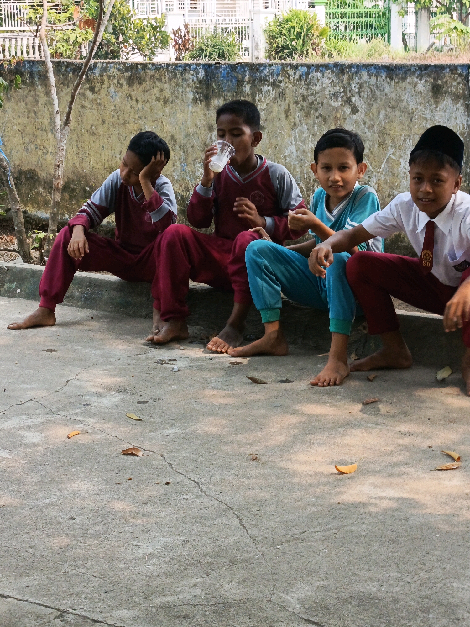 Four young boys are seated on a concrete ledge in what appears to be an outdoor schoolyard in Karya Dadi, Indonesia. The boys are wearing uniforms. From left to right, the first boy is wearing a maroon and gray long-sleeved shirt with maroon pants and has his hand on his head. The second boy is wearing a similar maroon and gray uniform and is drinking from a clear plastic cup. The third boy is wearing a teal short-sleeved shirt with blue pants. The fourth boy, on the far right, is wearing a white long-sleeved shirt, a red tie with a school emblem, and a black cap. He is also wearing maroon pants. All of the boys are barefoot. Behind them is a weathered, pale yellow wall with patches of blue and green, and beyond that, a white fence and lush green foliage. The ground in front of the ledge is a cracked concrete surface with scattered dry leaves. The lighting suggests it is daytime, with shadows cast on the ground.