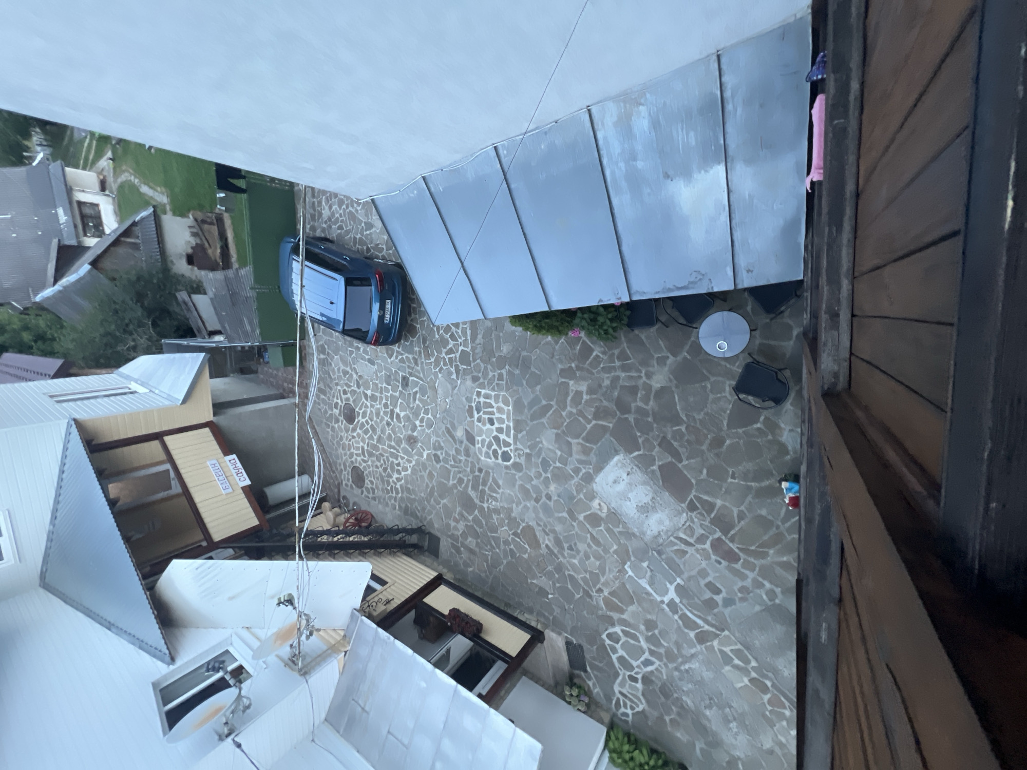 An aerial, high-angle, wide shot shows an outdoor courtyard with a blue SUV parked on a cobblestone surface. The courtyard is surrounded by buildings with white and beige exteriors and metal roofs. A sign with Cyrillic lettering is visible on one of the buildings. In the background, there are more houses and greenery. The foreground shows a wooden roof structure. The scene is located in Yaremche, Ukraine.