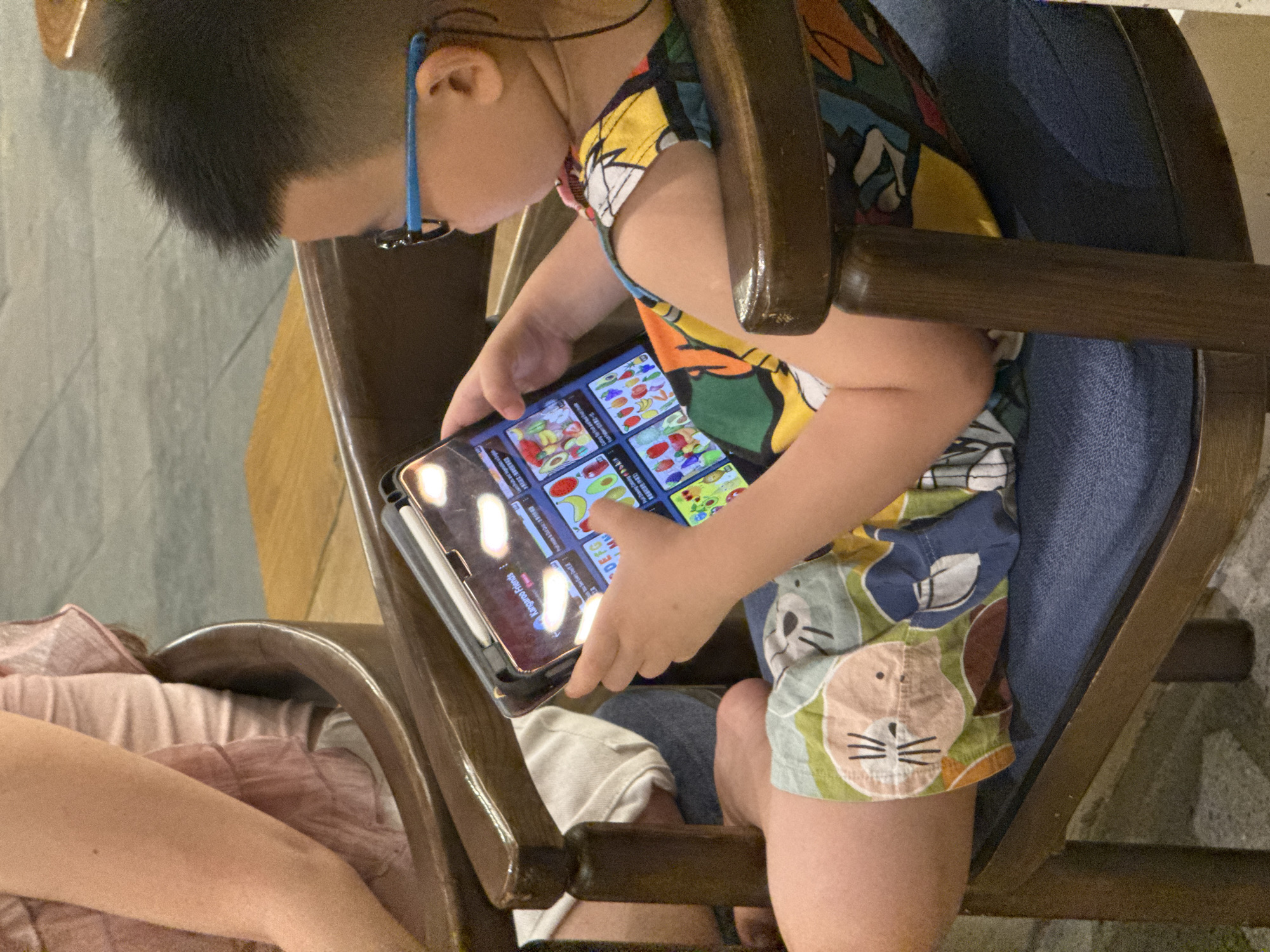 The image shows a young boy wearing glasses and a colorful patterned shirt sitting on a wooden chair with a blue cushion. He is holding a tablet device, which displays a game interface with various fruits and vegetables. A portion of an adult's arm, clad in a pink garment, is visible in the background. The setting appears to be indoors, possibly a cafe or restaurant, with wooden furniture and a tiled floor. The boy is focused on the tablet, engaged in playing a game. The scene captures a moment of leisure for the child.