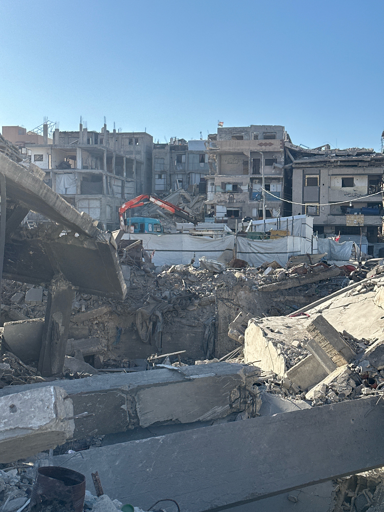The image depicts a scene of destruction in Gaza, likely a residential area. The foreground is filled with rubble, including large concrete slabs and smaller debris. In the mid-ground, a blue construction vehicle with an orange arm is visible amongst the debris and partially covered structures. Behind this, several partially collapsed buildings stand, their interiors exposed. Some buildings show signs of recent damage, with broken walls, exposed rebar, and incomplete construction. White tarpaulins are stretched over some areas, possibly for temporary shelter or to protect from the elements.
The sky is clear and blue, suggesting a sunny day, likely morning or afternoon given the strong shadows. The overall atmosphere is one of devastation and aftermath, with the destroyed buildings and scattered rubble indicating significant damage. A small flag is visible on top of one of the damaged buildings, but it is too small to discern its colors or insignia clearly. Some Arabic text is visible on the walls of the buildings, but it is not legible enough to be transcribed. The scene evokes a sense of loss and hardship.