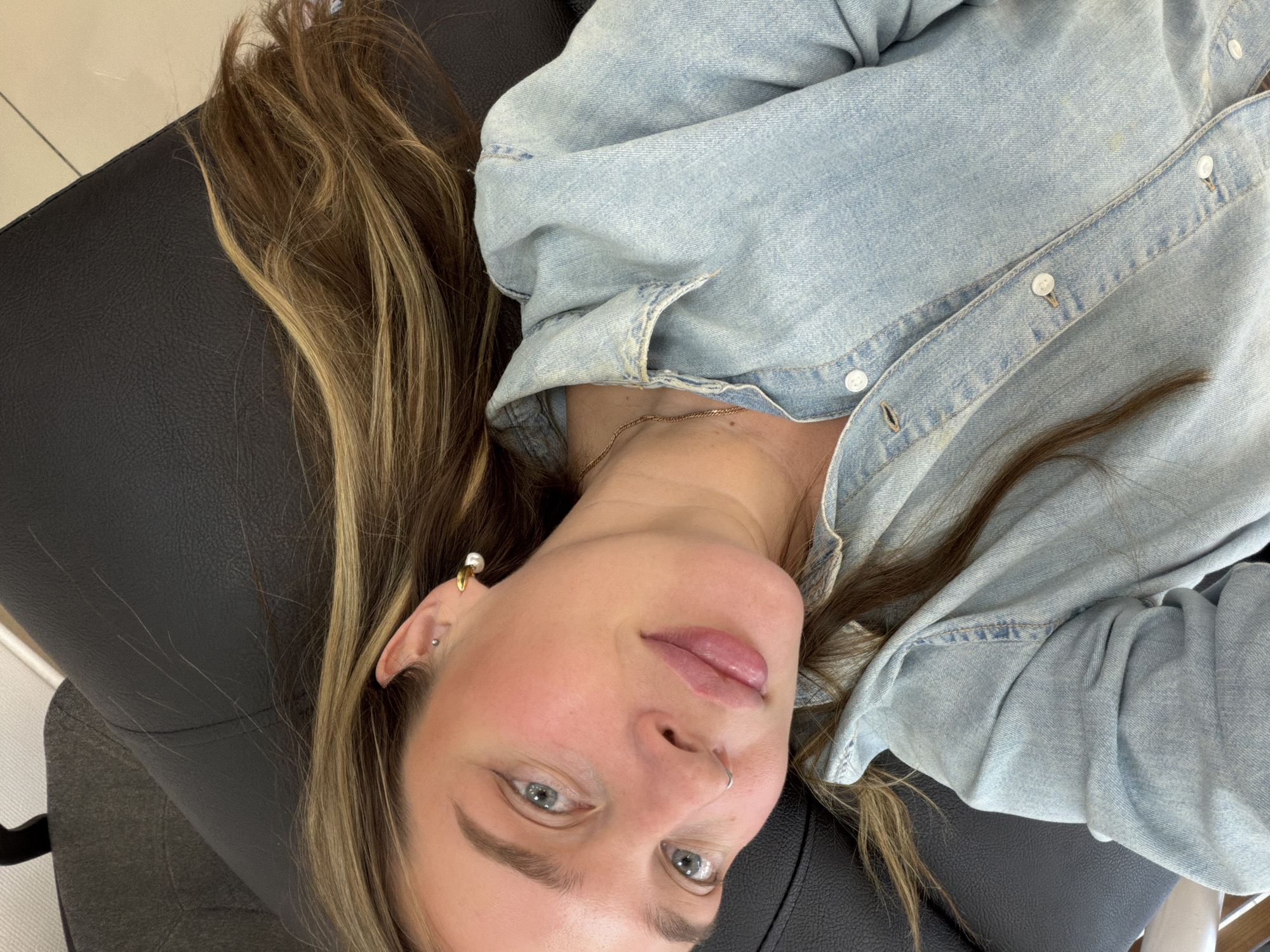 A woman with blonde-streaked brown hair, wearing a light blue denim shirt and gold necklace, is lying on a black leather surface, likely a chair or couch. She has blue eyes, a nose ring, and earrings. The location is unspecified and there is no indication of Chaikovsky, Russia.