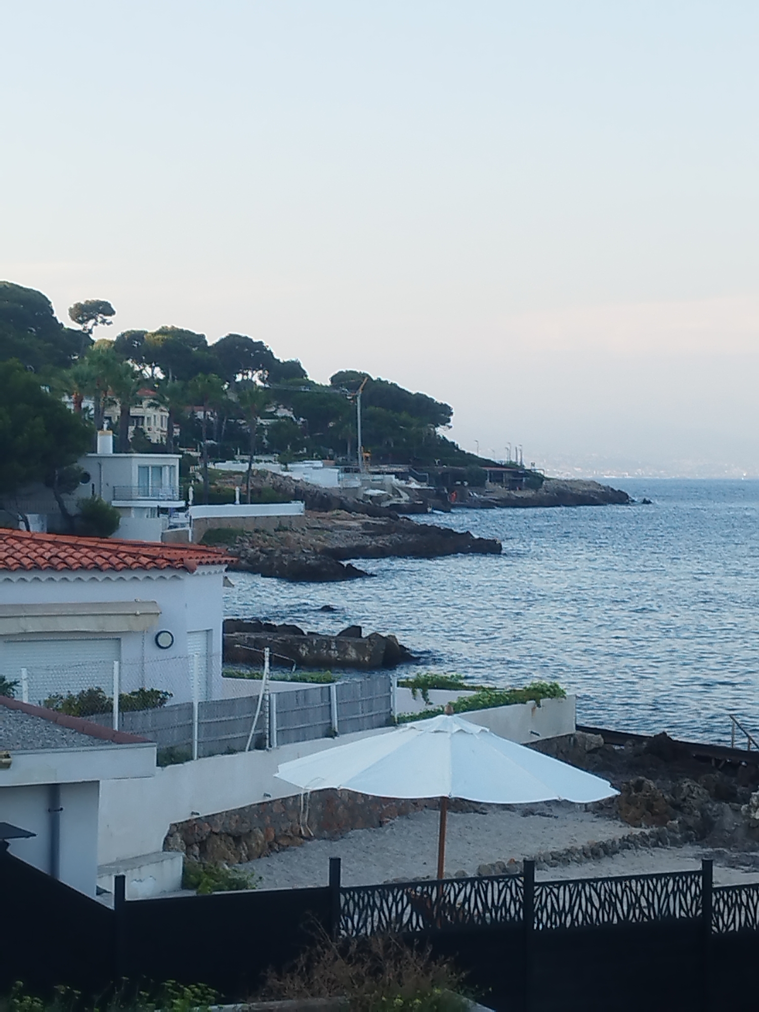 The image shows a coastal view of Antibes, France. In the foreground, there is a white umbrella on a sandy area with a dark metal fence. Behind it, there are buildings with white walls and red tile roofs, and a body of water that appears to be the Mediterranean Sea. The background features a rocky coastline with lush green vegetation and several houses nestled amongst the trees. The sky is clear and pale blue, suggesting it is either early morning or late afternoon. There are no people visible in the scene.