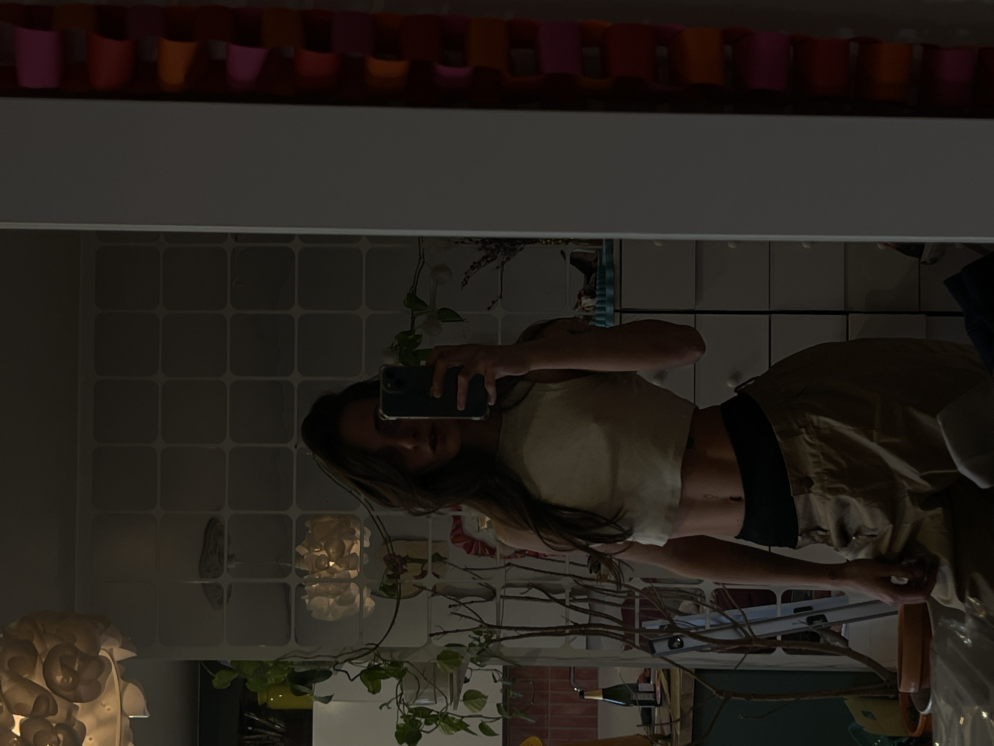 This image, originally rotated 90 degrees counter-clockwise, depicts a woman taking a mirror selfie indoors. The woman, who has long, dark hair, is reflected standing upright, holding a smartphone in front of her face, partially obscuring it. She is dressed in a light-colored, possibly cream or beige, crop top that exposes her toned midriff, paired with loose-fitting khaki or tan pants featuring a black elastic waistband.
The reflected background reveals an interior space, likely a room within a private residence. A wall covered with light grey or white square tiles is prominent. Various household objects and plants are visible: a line of small, brightly colored pots or cups (pink, orange, yellow, red) sits on a shelf above the tiled section. Several plants with green foliage and some with bare branches are arranged in the room, contributing to a natural or bohemian aesthetic. To the lower left, a distinctive decorative lamp or sculpture shaped like clustered white flowers emits a soft glow. Further to the right, a green bottle, resembling a wine or champagne bottle, rests on a surface. The ambient lighting is dim, indicating it is likely evening or nighttime. No discernible text is visible within the image. The location context provided is Krasnoyarsk, Russia.