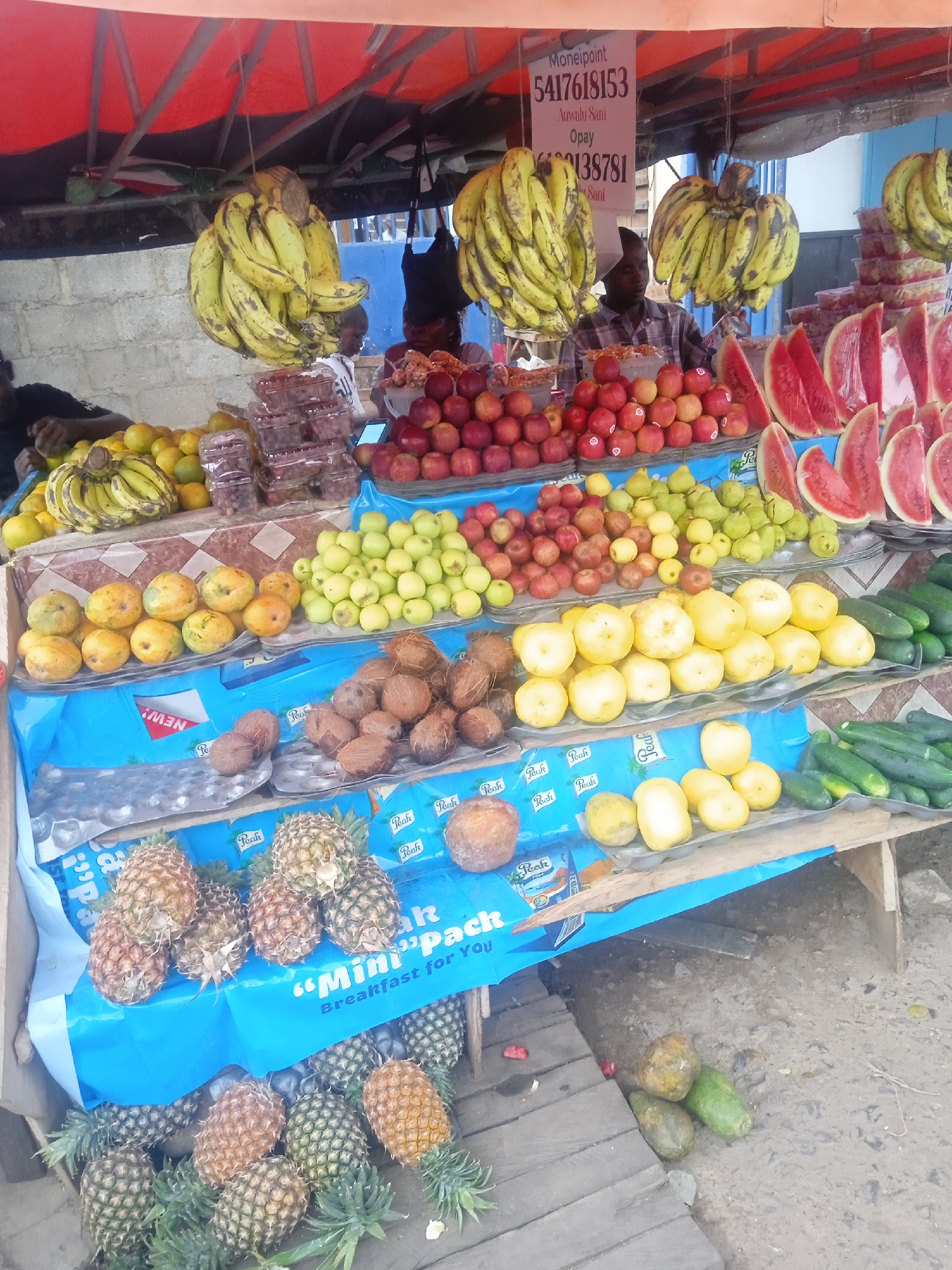 A fruit stand is in Jos, Nigeria. Bunches of bananas hang from the awning above. The stand has multiple tiers. From top to bottom, there are bananas, grapes, apples, and watermelon slices on the top tier. On the second tier, there are lemons, green apples, and red apples. The third tier has coconuts, tomatoes, and cucumbers. Pineapples are on the lowest tier. A sign displaying a phone number, likely for payment, is on the awning. There are people in the background.