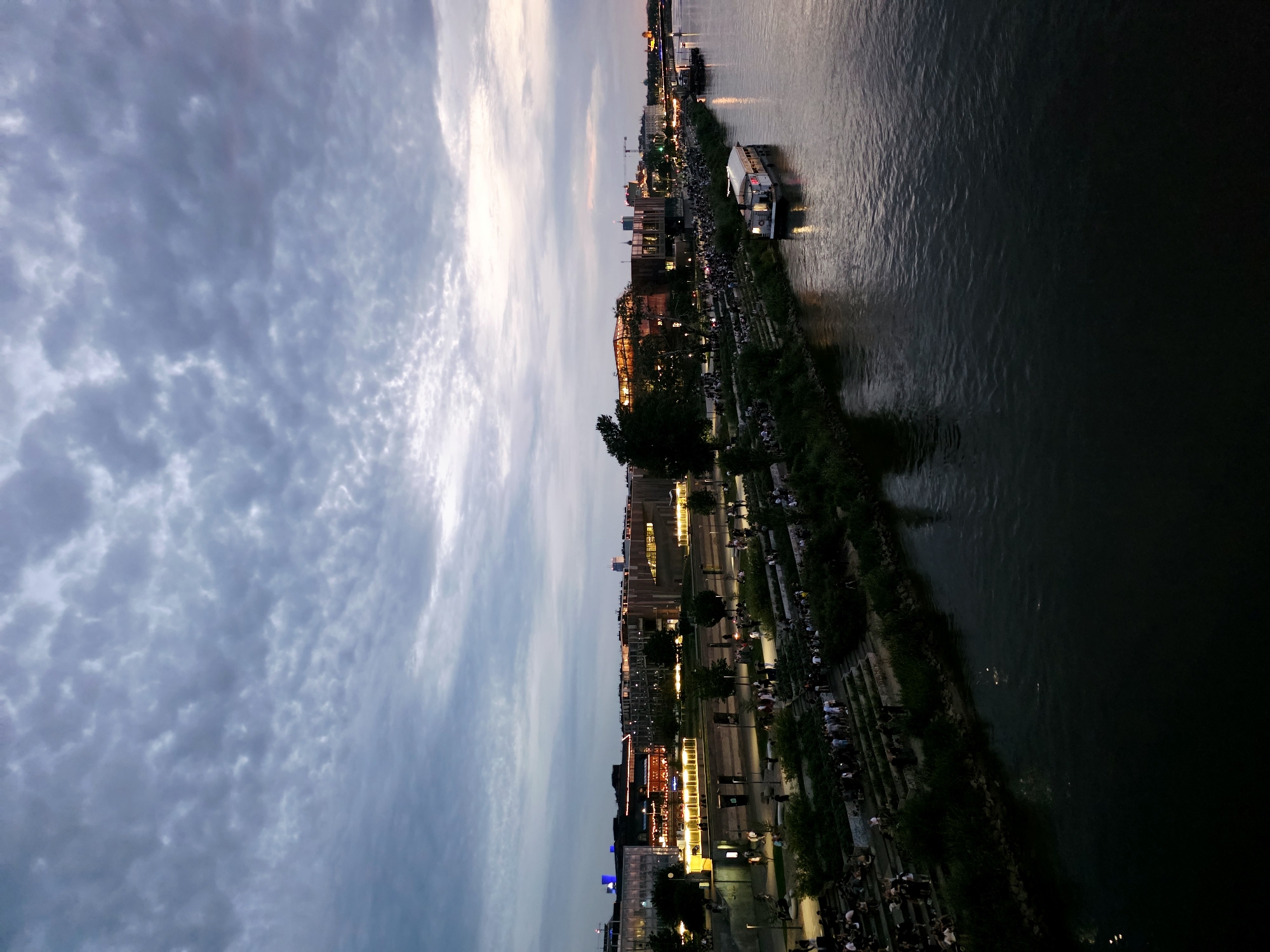 This is an eye-level shot of the Vistula River embankment in Warsaw, Poland, taken at dusk. The sky is filled with dramatic, textured clouds in shades of gray and blue, with a hint of warm light breaking through on the horizon. On the left bank of the river, a multi-level embankment is visible, densely populated with people sitting and standing on tiered steps and pathways. Lush green trees and shrubs line the embankment, interspersed with modern buildings that are illuminated with warm yellow lights. A white boat with a lit upper deck is moored in the dark, reflective waters of the river on the right side of the frame. The overall atmosphere is one of a vibrant urban evening scene.