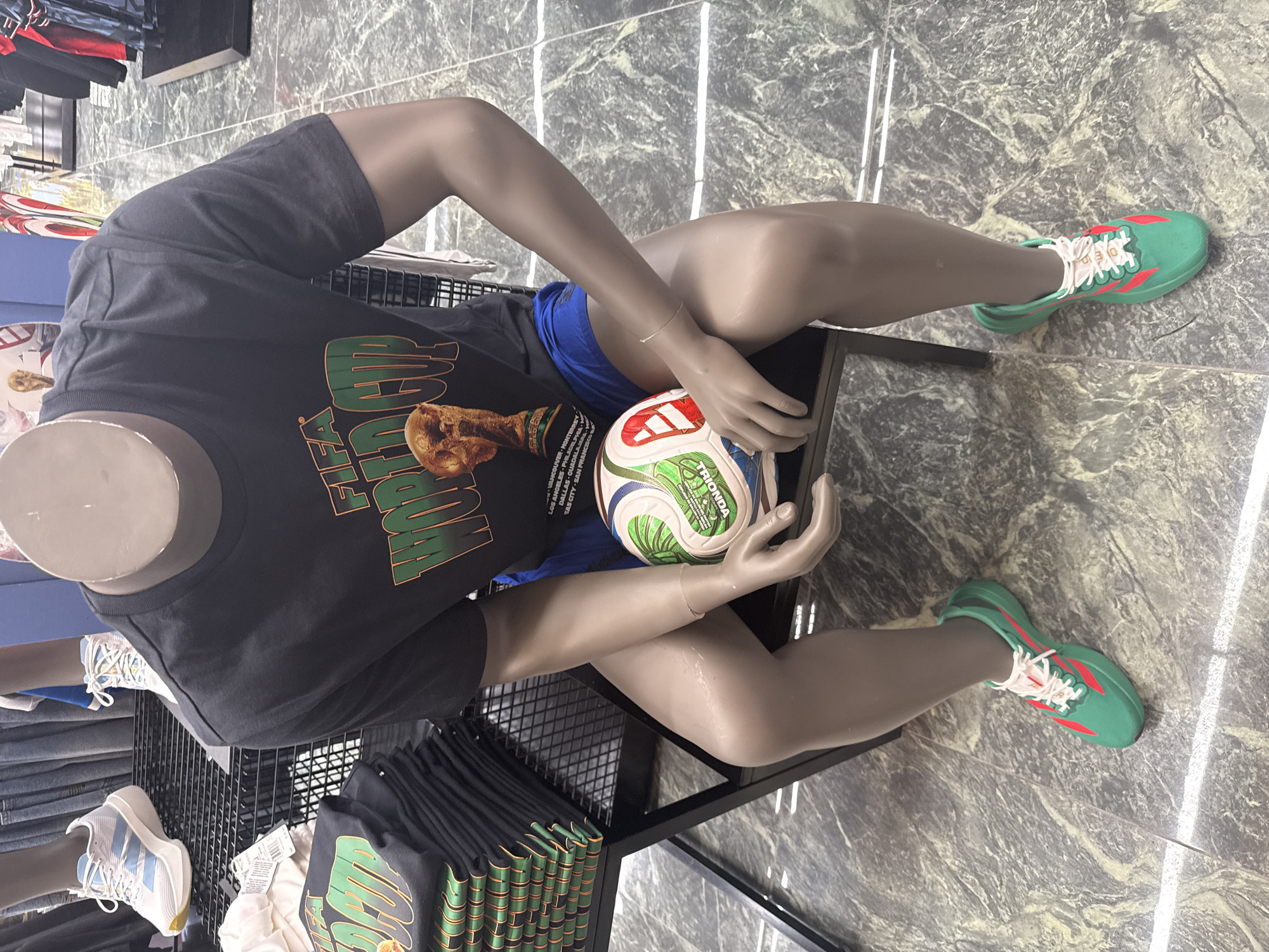This image presents a retail display within a commercial establishment, likely situated in Sunrise, United States of America. The central focus is a headless, dark-skinned male mannequin seated on a black metal chair or bench. The mannequin is dressed in a dark gray short-sleeved t-shirt featuring a prominent "FIFA WORLD CUP" graphic in green and orange, an image of the FIFA World Cup trophy, and a partially legible list of host cities for the 2026 event including Vancouver, Monterrey, Los Angeles, Guadalajara, Dallas, Houston, Kansas City, Atlanta, Miami, Philadelphia, Foxborough, Seattle, San Francisco, and New York/New Jersey. The mannequin also wears royal blue athletic shorts and emerald green and crimson red athletic sneakers with white laces. Its hands cradle a white, green, and blue soccer ball, branded "VOYAGIA" and "TRIOMPHE," in its lap. A thin, translucent bracelet is visible on the mannequin's right wrist.
To the left, a black metal grid display unit holds several neatly stacked black t-shirts, identical to the one worn by the mannequin, with a white price tag attached to the topmost stack. In the background, on a similar black metal grid shelf, the lower legs and feet of two additional mannequins are visible, each displaying athletic shoes. One wears white sneakers with light blue Adidas stripes, and the other wears white sneakers with yellow laces. The flooring is composed of gray and white marbled tiles with visible grout lines, extending throughout the visible area of the store. Additional retail fixtures and blurred apparel are discernible in the deeper background.