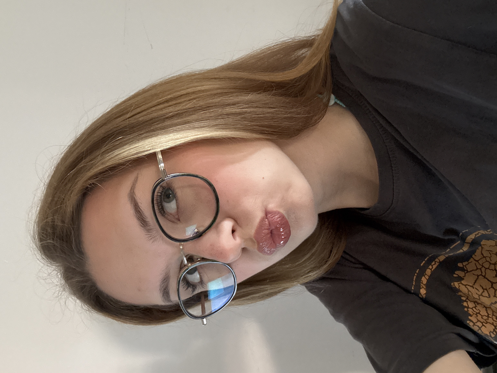 A young woman with light brown hair and wearing glasses is positioned at an angle in the image. She has a light complexion and is making a kissing face. She is wearing a dark-colored t-shirt with a design on it. The background appears to be a plain, light-colored wall. The image provides no information about the location being in Kragujevac, Serbia. The scene is a close-up self-portrait.