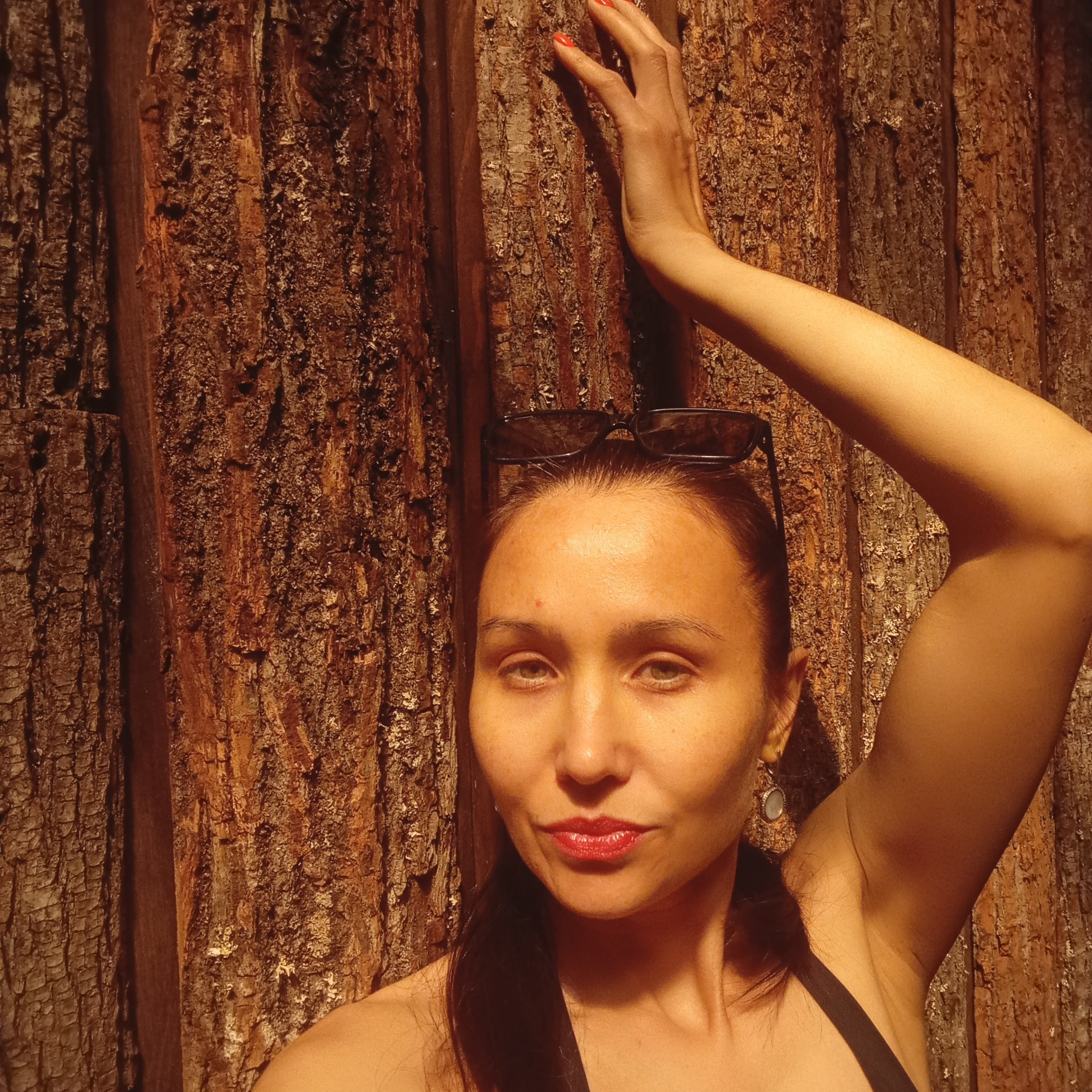 A woman stands in front of a textured wooden wall, her right arm raised and hand touching the wall, her left arm by her side. She has dark hair pulled back, sunglasses resting on her head, and wears earrings. She faces the camera with a slight smile, wearing lipstick. The lighting suggests an outdoor setting with strong sunlight. The location is Izhevsk, Russia.