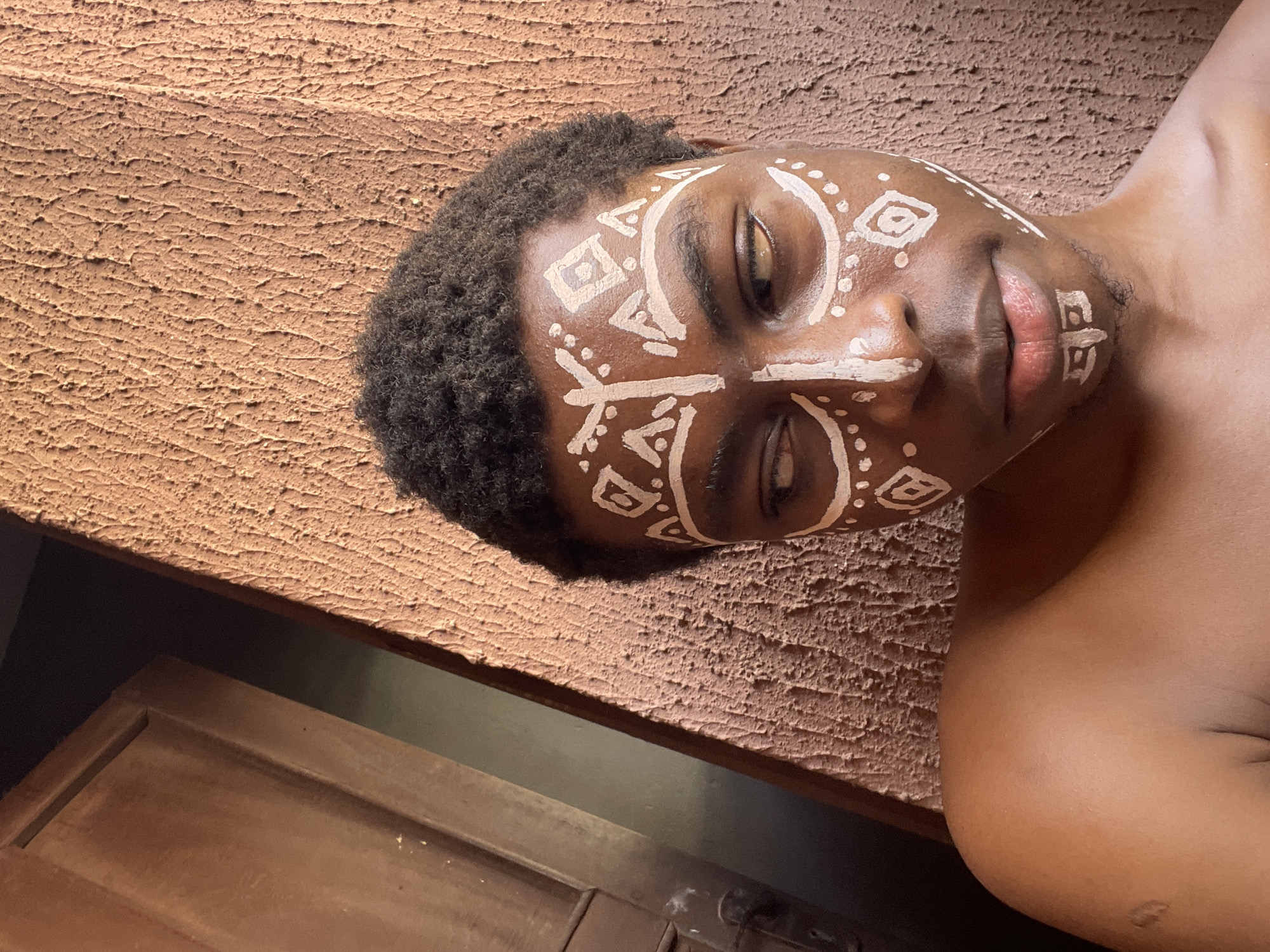 A horizontally-oriented close-up depicts a person lying down, facing the camera. The individual has dark skin, short, curly black hair, and their face is adorned with white tribal-style paint. Their eyes are mostly closed. The person is positioned against a textured brown surface. Part of a wooden door is visible in the lower-left corner of the image. The location within Nkpor, Nigeria is not discernible based solely on the image.