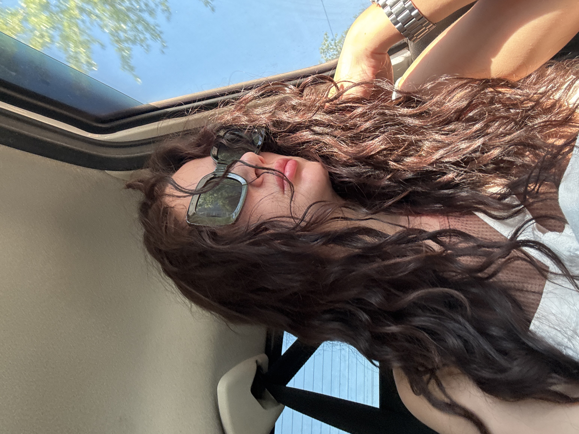 A woman with long, curly brown hair is lying down in a car with an open sunroof. She is wearing sunglasses. Her face is partially visible. One arm is raised towards the opening of the sunroof; a silver watch is visible on her wrist. A seatbelt is visible next to her shoulder. Through the sunroof, a blue sky and foliage are visible. Vertical blue metal is visible through the car window. The location is unknown, but not necessarily Chaikovsky, Russia.