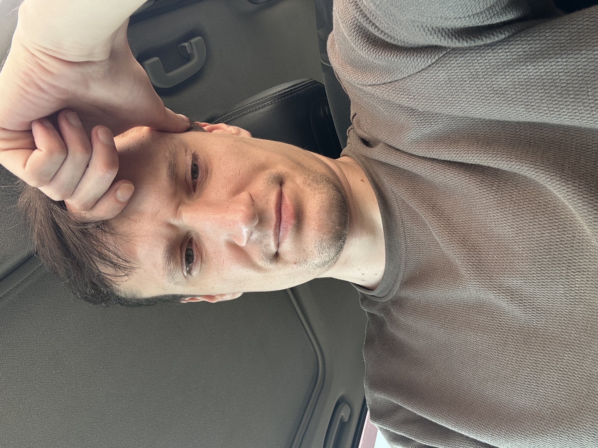 A man with brown hair and stubble is leaning his forehead against his hand while inside a car. He is wearing a brown, waffle-knit shirt. The car interior is visible, including the headliner and a portion of a black leather seat. The man's expression appears tired or thoughtful. The image was taken in Shatava, Ukraine.