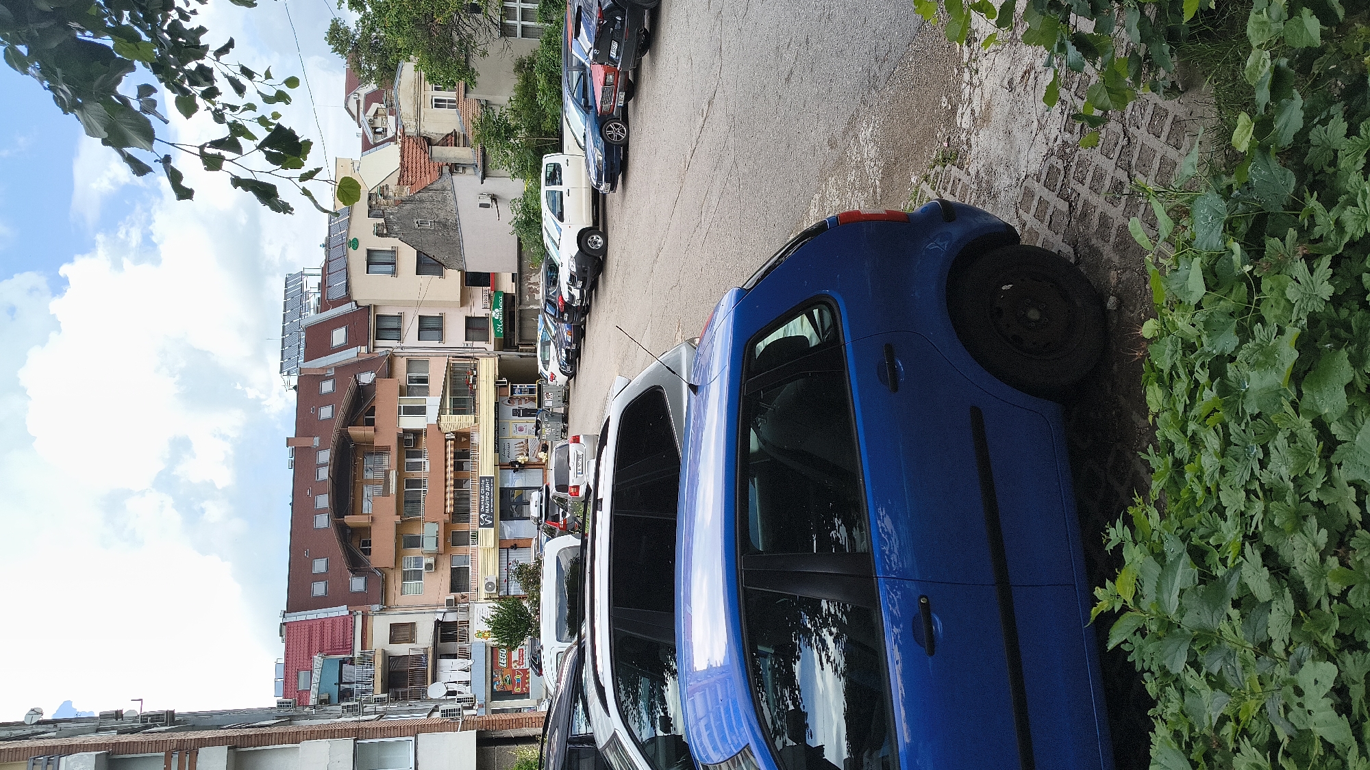 This image depicts an urban scene in the city of Shumen, Bulgaria, featuring a parking lot bordered by various multi-story buildings under a partly cloudy sky.
In the immediate foreground, dense green foliage, consistent with bushes and ground cover, occupies the bottom portion of the frame, obscuring the lower half of the nearest vehicle. The closest vehicle is a blue four-door hatchback, oriented with its passenger side visible. Behind it, a silver five-door SUV is parked. The parking lot surface consists of worn asphalt or concrete, with sections of interlocking paver blocks visible in the foreground and cracks where vegetation is starting to emerge.
Further into the parking lot, multiple cars are visible, including a white pickup truck parked horizontally, followed by a row of diverse passenger vehicles. One silver car in the mid-ground displays a Bulgarian license plate "CA 9467 HT". No individuals are visible anywhere in the scene.
The background is dominated by a collection of residential and commercial buildings. On the far left, a tall, light-colored building with multiple balconies and external air conditioning units is partially visible. The central background features a row of buildings with varying architectural styles and colors, ranging from pinkish to beige and light yellow. Most have prominent red or brown tiled roofs, many with dormer windows and skylights. One central building exhibits a distinctive curved balcony structure. At ground level, several businesses are identifiable by their signs; a yellow sign with "LEGO" is visible, along with a white sign displaying "МАЙТРО ДЕНТ" (MAITRO DENT), indicating a dental practice. A green awning further right reads "Меденка" (Medenka). To the far right, an older, possibly residential, building with a more traditional orange tiled roof and a distinct facade stands next to the more modern structures. Overhanging green tree branches partially frame the upper right portion of the image. The sky is bright with a mix of white clouds and blue patches.