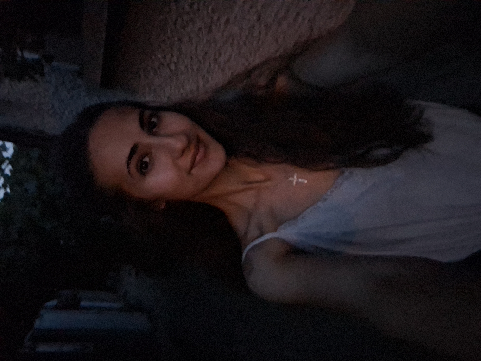 A young woman with long, dark hair is taking a selfie outdoors at dusk. She is wearing a white camisole with lace trim and a silver cross pendant. Her face is turned towards the camera, and she has a slight smile. The background is dark, with some suggestion of buildings and foliage. The lighting is dim, creating a moody atmosphere. The scene appears to be in a quiet, possibly residential, area.