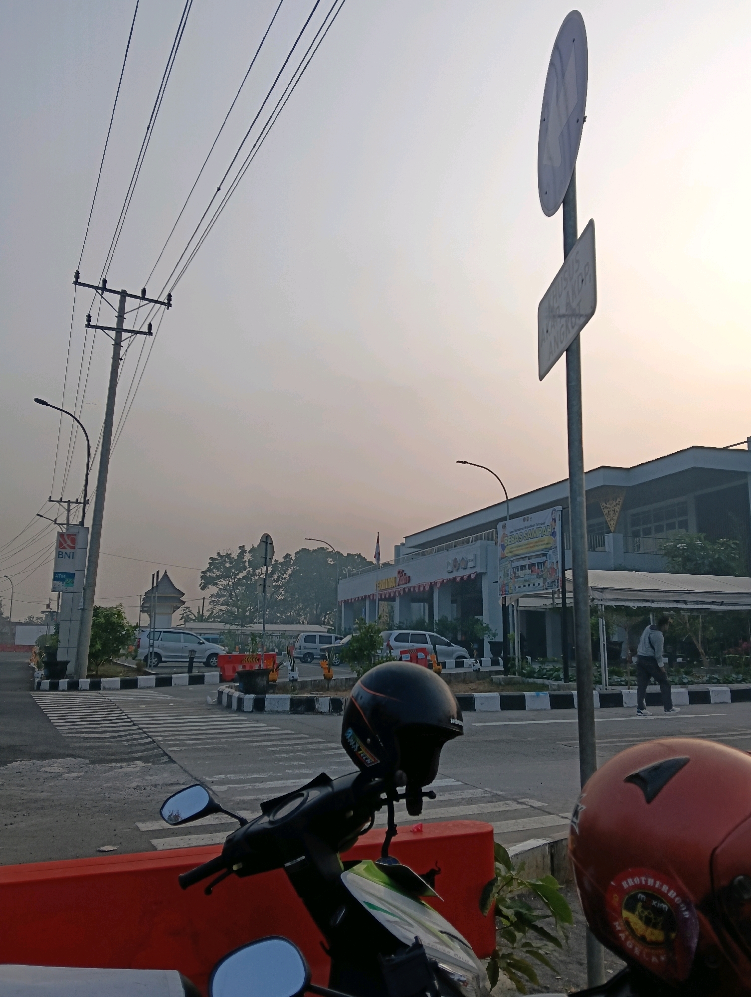 The image captures a street scene in Magelang, Indonesia, characterized by a hazy, overcast sky and an atmosphere of early morning or late afternoon. In the foreground, two motorcycles are parked, one with a black helmet and the other with a red helmet bearing a "Brotherhood M.XIM MAGelang" sticker. A red barrier separates the foreground from the street, which features a pedestrian crossing with striped markings.
Behind the barrier, several cars are visible, including a silver sedan and two vans. The street is lined with trees and a building that appears to be a commercial establishment with signage that includes the text "BEBAS SAMPAH" (meaning "No Trash") and graphics depicting a clean environment. A utility pole with electrical wires stands prominently to the left of the frame, along with a sign for BNI (Bank Negara Indonesia) and an ATM.
Further in the background, another signpost with a circular and rectangular sign is present. The overall environment suggests a public area within the city, possibly near a shopping center or a transit point. The light conditions indicate low visibility, likely due to mist or pollution.