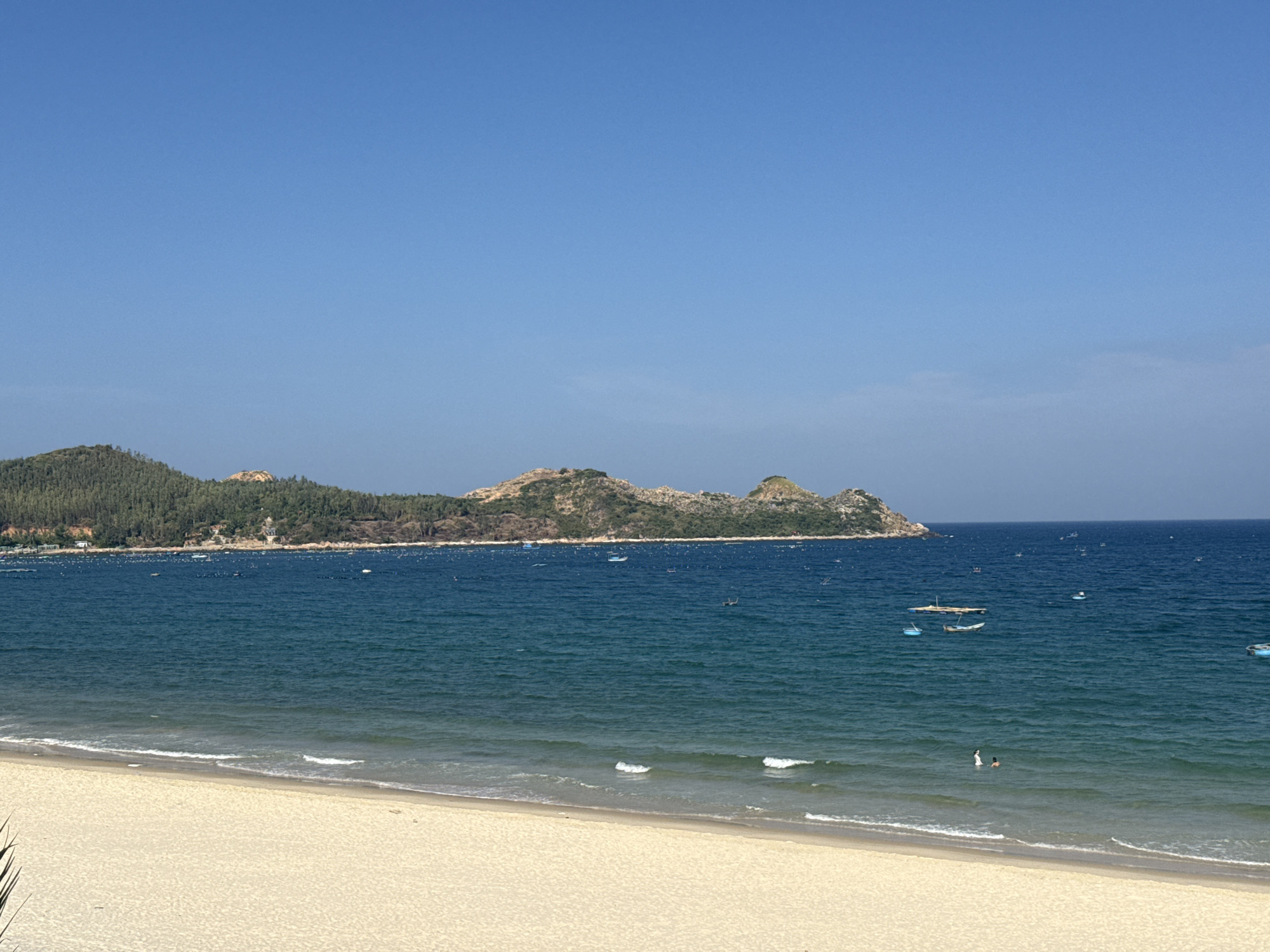 The image depicts a serene coastal scene with a clear blue sky overhead. In the foreground, a sandy beach stretches out, meeting the gentle waves of the ocean. The water is a deep blue, with white crests of waves breaking near the shore. Small boats are scattered across the sea, and a few people are visible in the water, swimming or wading. In the distance, a lush, green, mountainous landscape rises from the coastline, with trees covering its slopes. The overall impression is one of a tranquil day at the beach, with nature taking center stage.