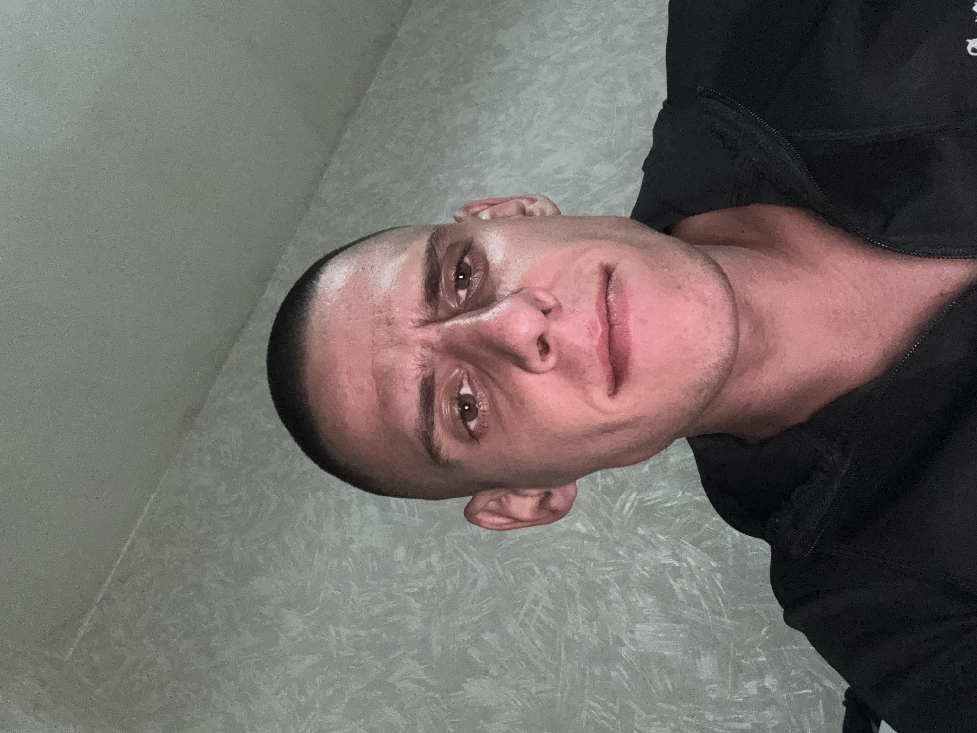 A man with a shaved head is posing for a selfie. He is wearing a black zip-up hooded sweatshirt. The background is a light-gray wall. There is no indication this image was taken in Sofia, Bulgaria.
