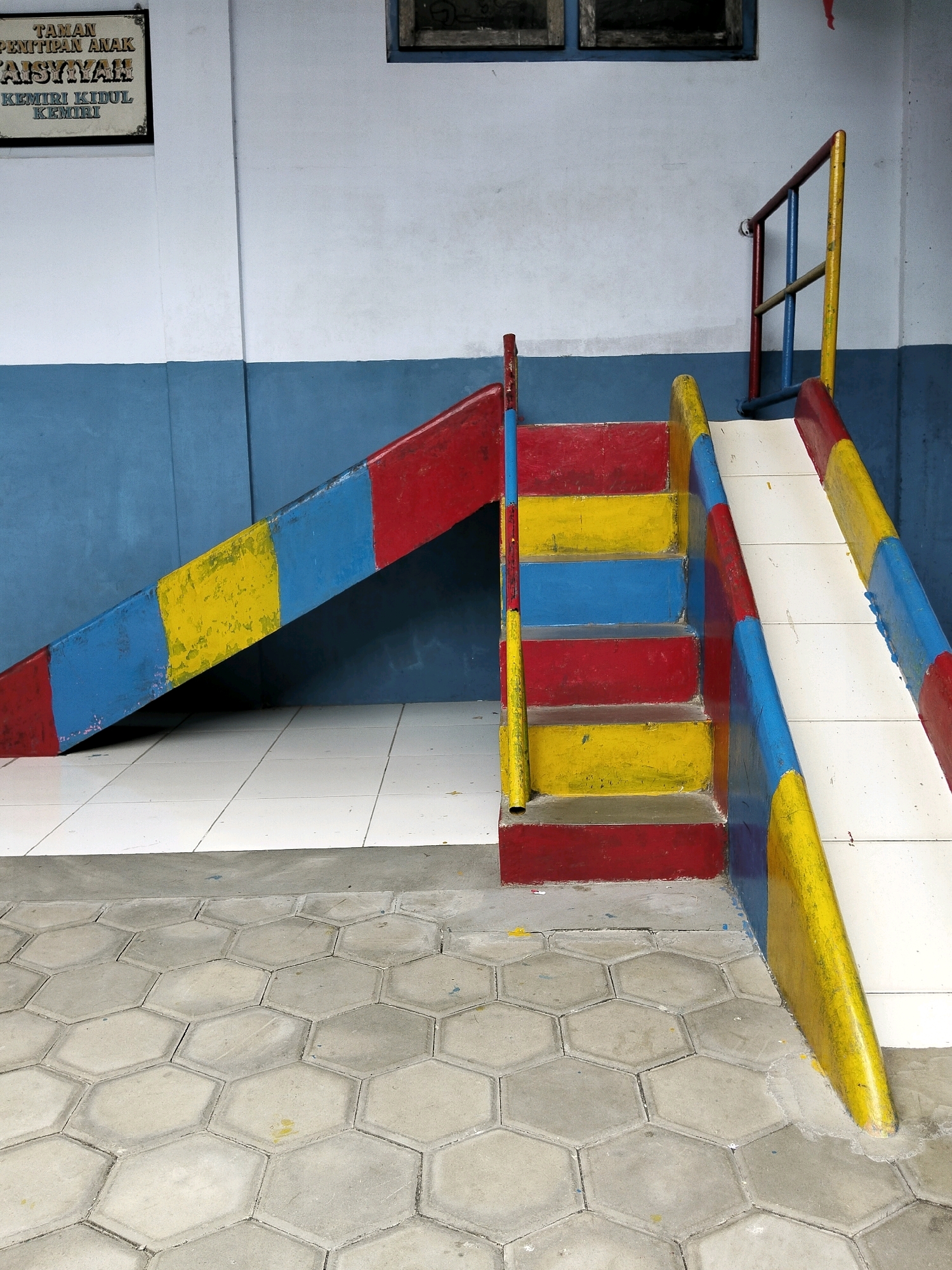 The image shows a brightly colored children's slide and stairs set against a blue and white wall. The slide is painted in red, yellow, and blue sections. The stairs are also multi-colored with red, yellow, and blue steps. A metal railing, painted red and yellow, is attached to the side of the slide structure. The ground in front of the slide is covered in hexagonal gray paving stones, and behind the slide, white tiled flooring is visible. A sign is mounted on the wall above the slide, with text in Indonesian that translates to "Day Care Center AISYIYAH, KEMIRI KIDUL KEMIRI." This indicates the location is likely a day care facility in Kemiri Kidul, part of Krajan Lor, Indonesia. The overall scene depicts an outdoor play area.