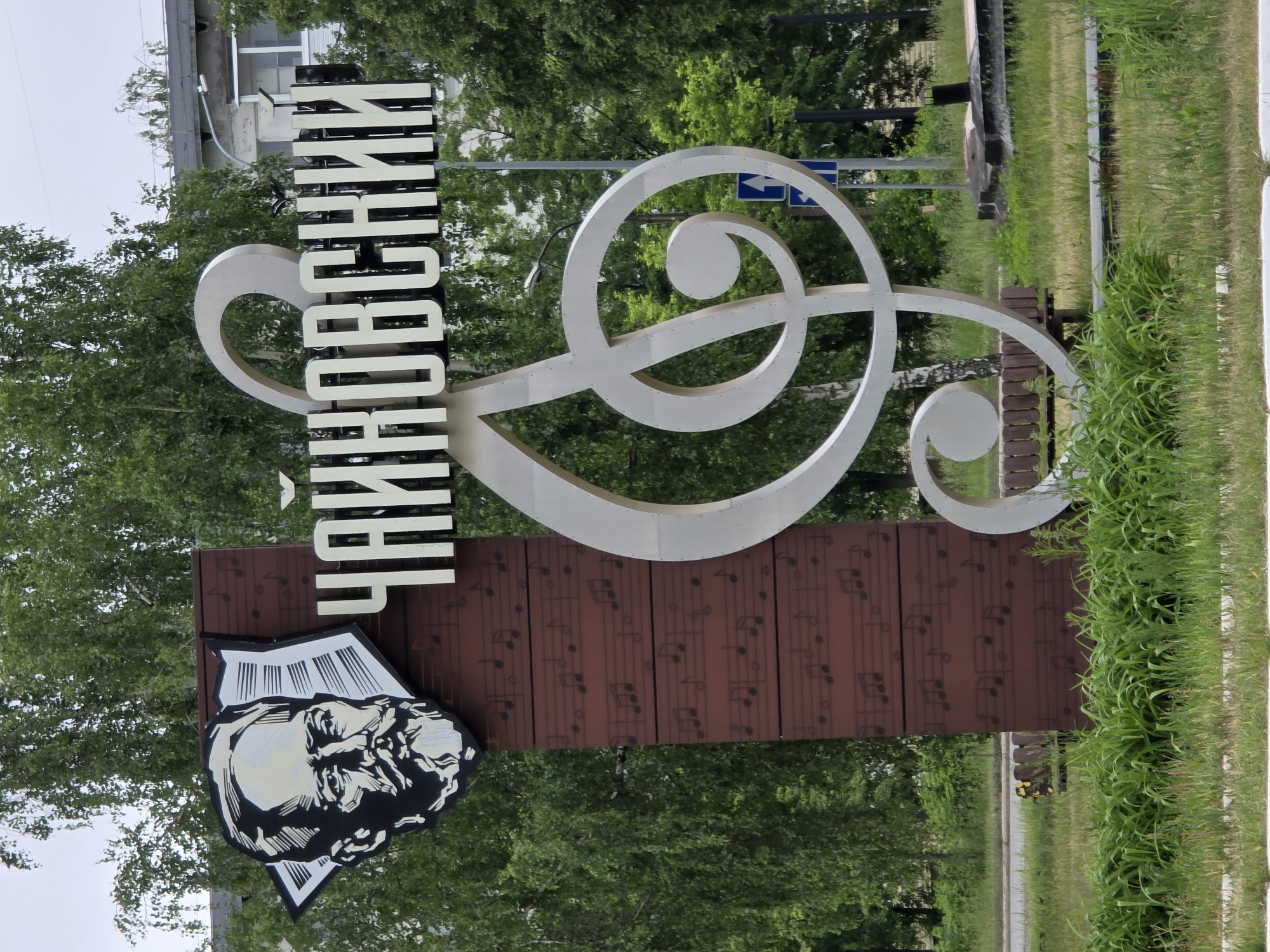 The image shows a vertical sign in the city of Chaikovsky, Russia. The sign has a large metal treble clef at the top, and below that, the name "Chaikovsky" is written vertically in Cyrillic letters. Below the name is a rectangular board with musical notes printed on it, and below that, an image of a man's face is displayed on a star-shaped background. The sign is surrounded by greenery, including trees and grass. There are buildings and street signs visible in the background.