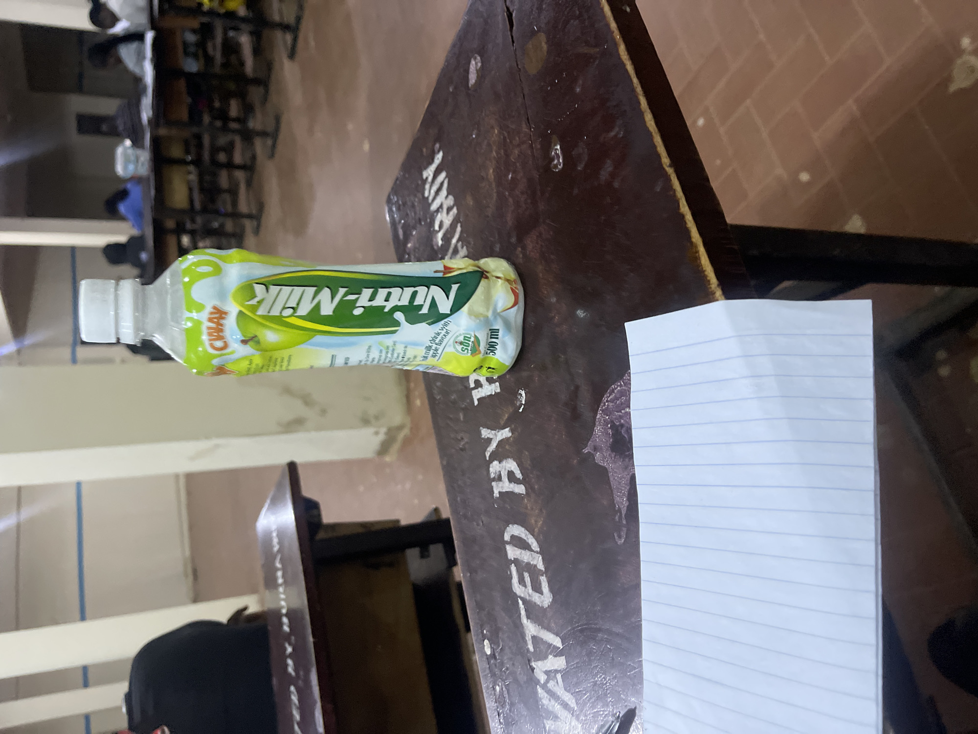 A bottle of Nutri-Milk with an apple flavor is placed on a wooden desk in what appears to be a classroom. The desk has faded white lettering that reads "VATED BY BURNA W... BOY." A sheet of lined paper is placed in the foreground. In the background, students are seated at desks, facing forward. The floor is tiled with red bricks. The overall scene suggests a school environment, likely during a lesson or study period, possibly in Rafin Par, Nigeria, given the context provided.