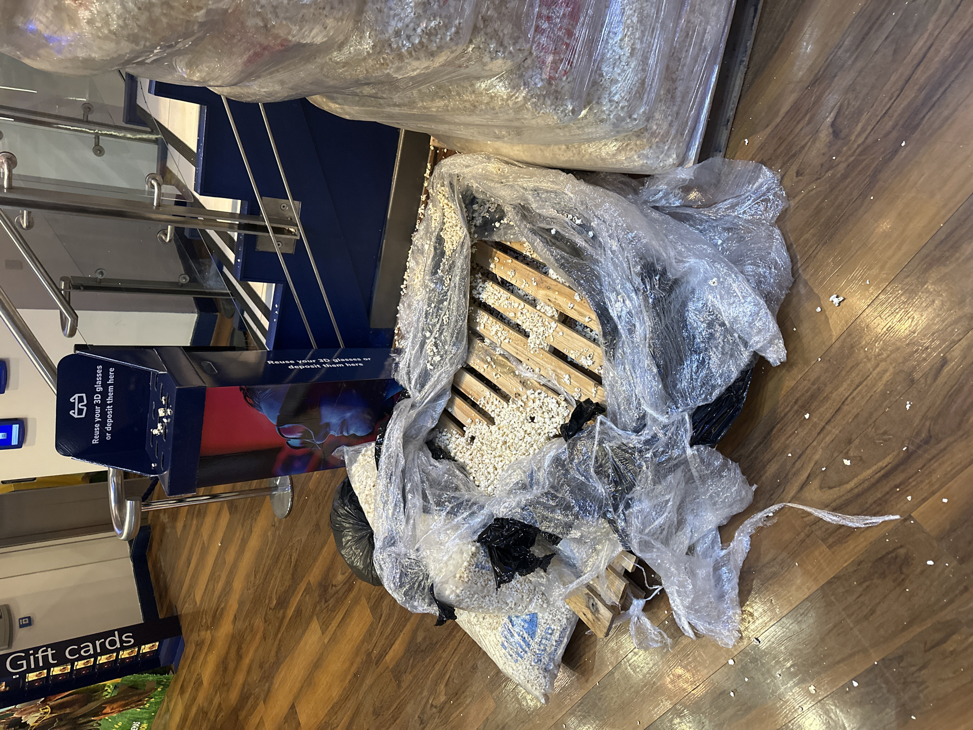 The image shows a pile of what appears to be spilled or discarded popcorn in a commercial setting. There are bags of popcorn, some broken open, spilling onto a wooden pallet and the surrounding floor. Clear plastic wrapping or bags are draped over the pile. Nearby is a receptacle labeled "Reuse your 3D glasses or deposit them here," suggesting a movie theater or entertainment venue. In the background, there's a set of stairs with a blue and silver railing. A "Gift Cards" display is partially visible in the lower left corner. The location is indicated as Tonbridge and Malling, United Kingdom. The floor has a wood-grain pattern.