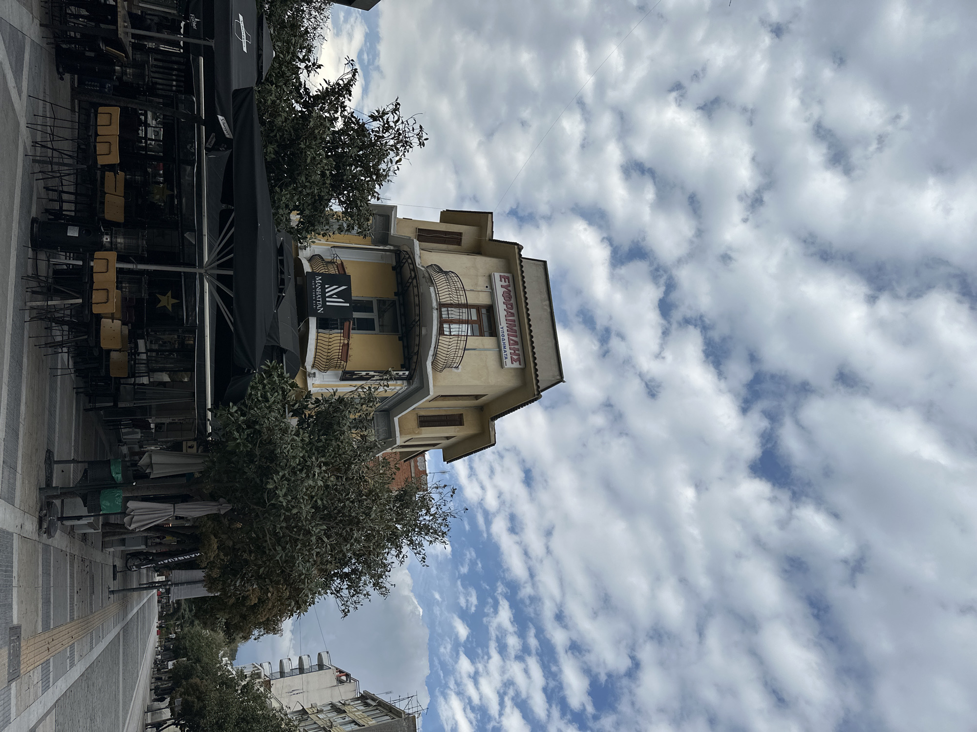 The image shows a street scene in Serres, Greece. The view is framed by a blue and white sky filled with clouds, a street to the left and a building in the center. The street is lined with trees and includes tables and chairs, some stacked. The building, which appears to be three stories tall, features a sign on its upper portion. The signage reads ΕΥΘΡΑΙΜΙΑΔΗΣ, ΥΠΟΔΗΜΑΤΑ in Greek letters. The side of the building features a sign for the Manhattan bar. Balconies with ornate railings adorn the building.