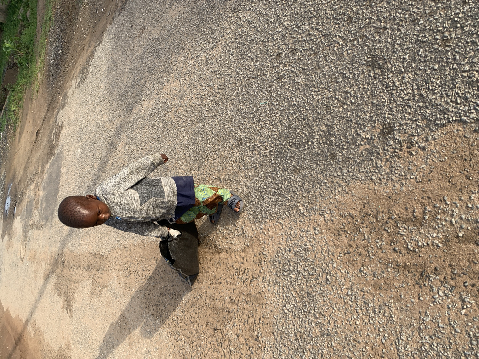 A young boy stands on a gravel and sandy surface, likely a roadside or path, partially adjacent to a paved area and some vegetation. The boy is wearing a grey hooded sweatshirt, a patterned fabric around his waist, and blue sandals. The scene is sunlit, casting shadows. The background is dominated by the textured gravel surface. The boy is the only visible person in the frame. It is impossible to ascertain the precise location in Abuja based solely on this image.