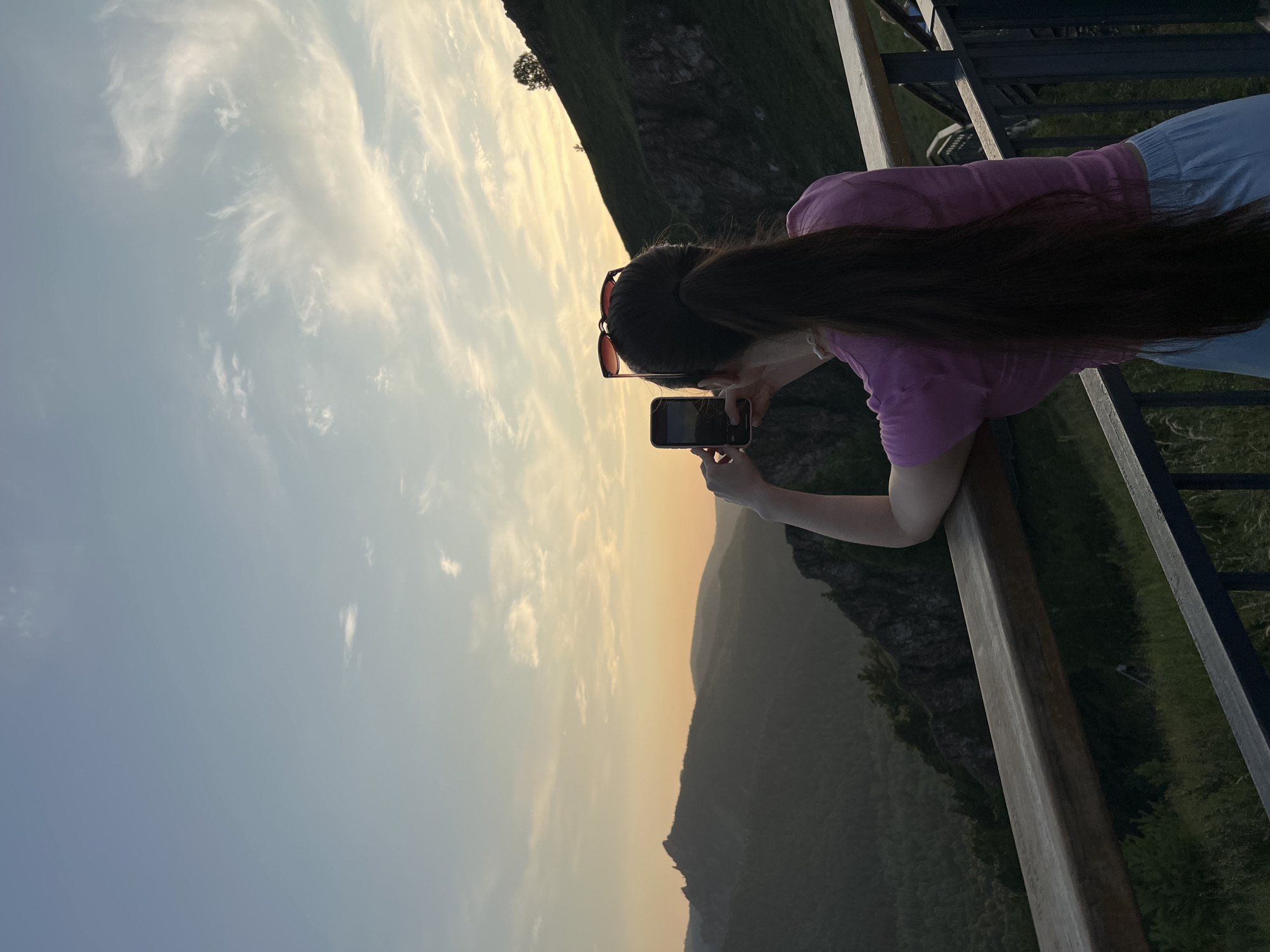 A woman with long dark hair pulled back in a ponytail is standing on a wooden railing, taking a photo with her mobile phone of a mountainous landscape at sunset. She is wearing a pink t-shirt and light blue shorts. Her sunglasses are perched on top of her head. The sky is a soft blend of blue, yellow, and orange, with wispy clouds. The mountains are layered in the background, with trees covering their slopes. The scene is likely from a vantage point overlooking the Yenisei River in Krasnoyarsk, Russia, given the characteristic topography of the area, such as the Stolby Nature Reserve.